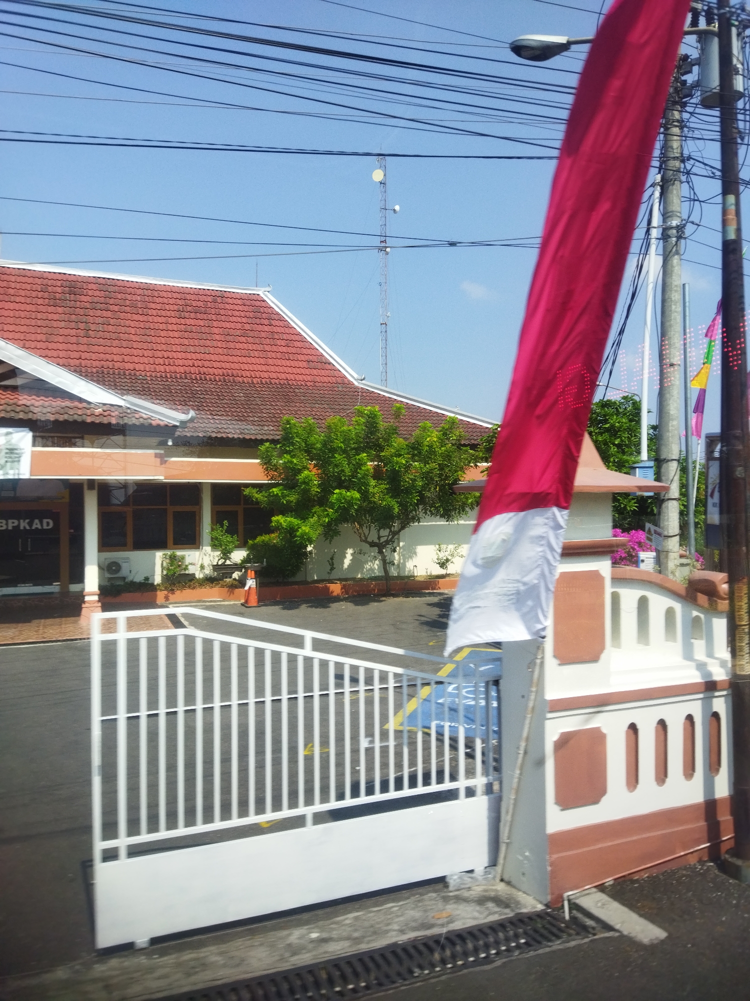 This is a medium-angle, outdoor shot of a building entrance with a white metal gate in the foreground. The building has a red tile roof and is painted white and tan. A large red and white flag, likely the Indonesian flag, is prominently displayed to the right, partially obscuring the view of the building and a utility pole. The gate is open, revealing a paved driveway or courtyard area. Beyond the building, trees and a communication tower are visible against a clear blue sky. The scene suggests a civic building or office, possibly in a public space within Wonokarto, Indonesia, given the presence of the Indonesian flag.