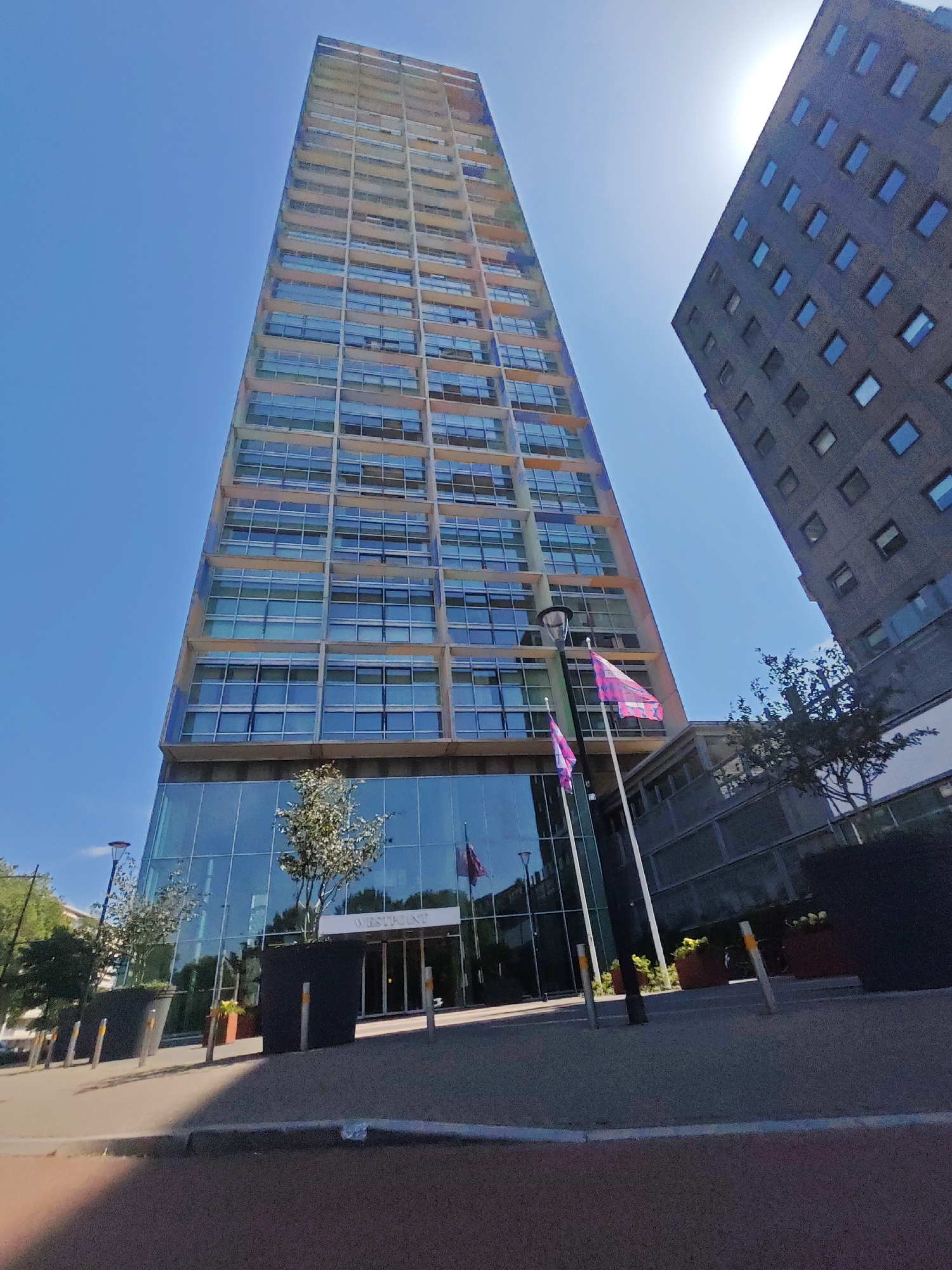 A tall, slender skyscraper with a facade of glass and beige concrete panels dominates the image, angled upwards against a clear blue sky. To the right, another, shorter building with dark grey, textured concrete and a grid of small, blue-tinted windows rises, partially obscuring the sky. Two flags, predominantly pink with a white design, fly from poles in front of the skyscraper's glass entrance. The building's name, "WESTPOINT", is visible above the entrance. Planters with small trees and shrubs are placed along the paved area in front of the building. The scene appears to be a contemporary urban setting in Tilburg, Netherlands, on a bright, sunny day.