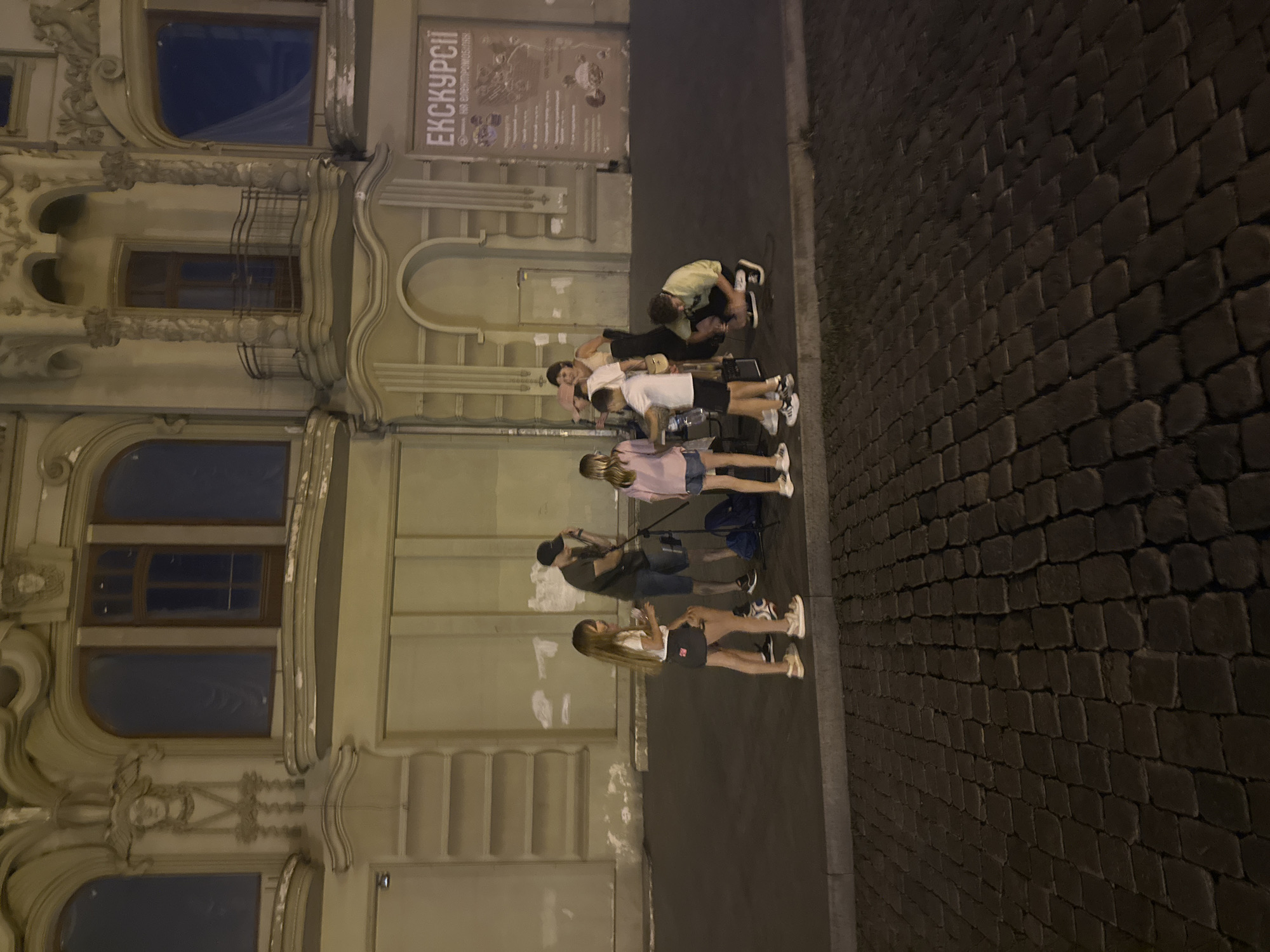 In Odesa, Ukraine, on a cobblestone street at dusk, a group of people are gathered in front of an ornate building. The building features classical architectural elements such as columns, arches, and decorative moldings. A large poster advertising "EXCURSIONS" is displayed on the building's facade.
A man wearing a black cap and dark shirt stands near a tripod with audio equipment. Three young girls, two in casual wear and one in a white shirt and dark skirt, are positioned around a table with musical instruments, possibly drums. Another young boy in a white t-shirt and black shorts is also near the instruments. A woman in a light-colored top and a black skirt stands behind the children. A young man in a light green shirt is crouched down, appearing to be setting up or adjusting equipment. The scene suggests a street performance or practice session is taking place.