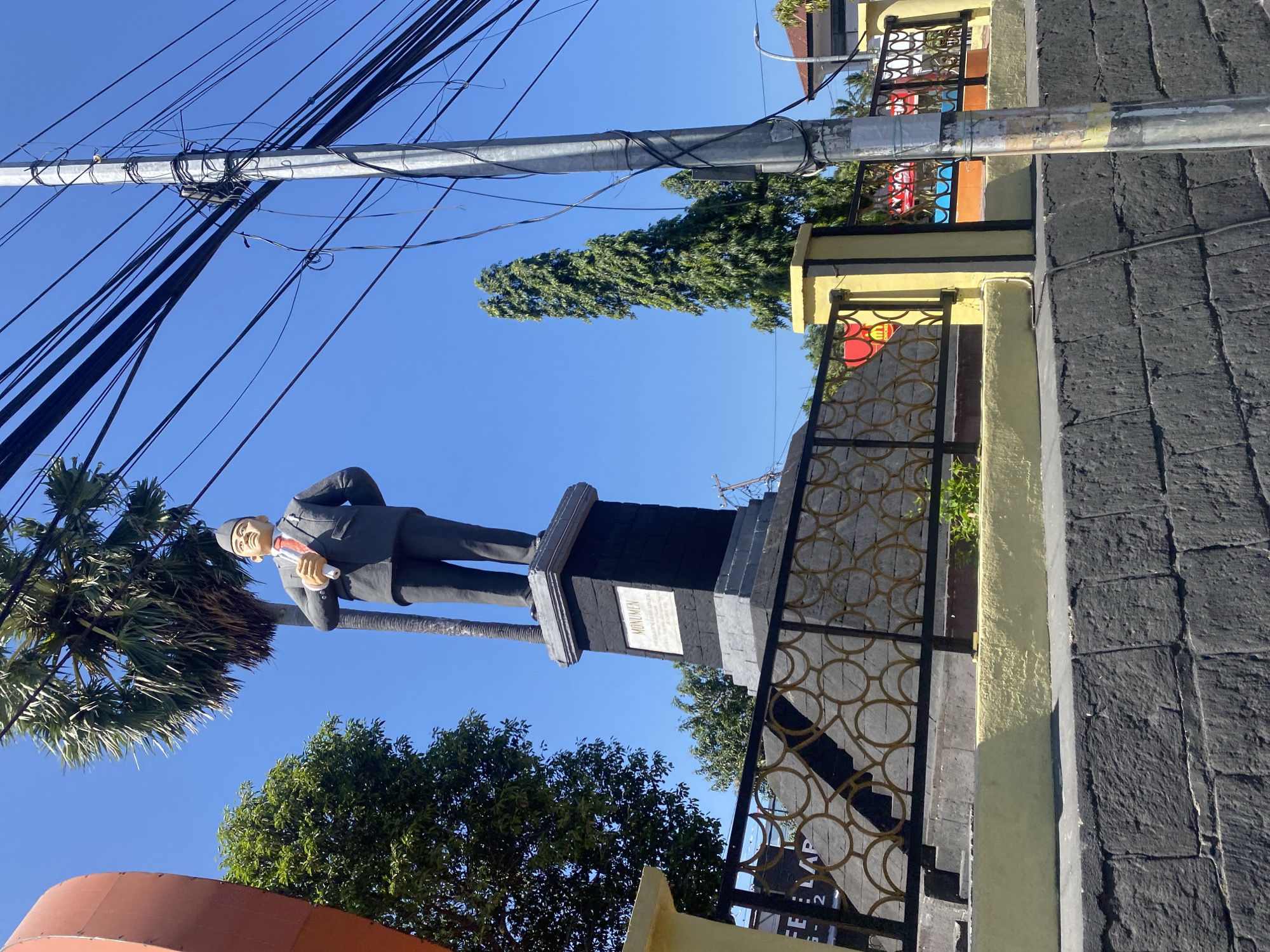 The image shows a statue of a man in a suit and tie, standing on a pedestal with the word "MONUMEN" inscribed on it. The statue is located outdoors, with a clear blue sky and some trees in the background. A utility pole with many cables is visible to the right of the statue. A fence with a decorative pattern runs in front of the statue, separating it from the foreground. There is a wall made of dark, rough-hewn stones in the immediate foreground. Based on the presence of the "MONUMEN" inscription and the context of the city of Bantaeng, Indonesia, this is likely a monument in the city, possibly a historical figure or a significant person in the region's history. The overall scene depicts an outdoor public space with a monument as the central subject.
