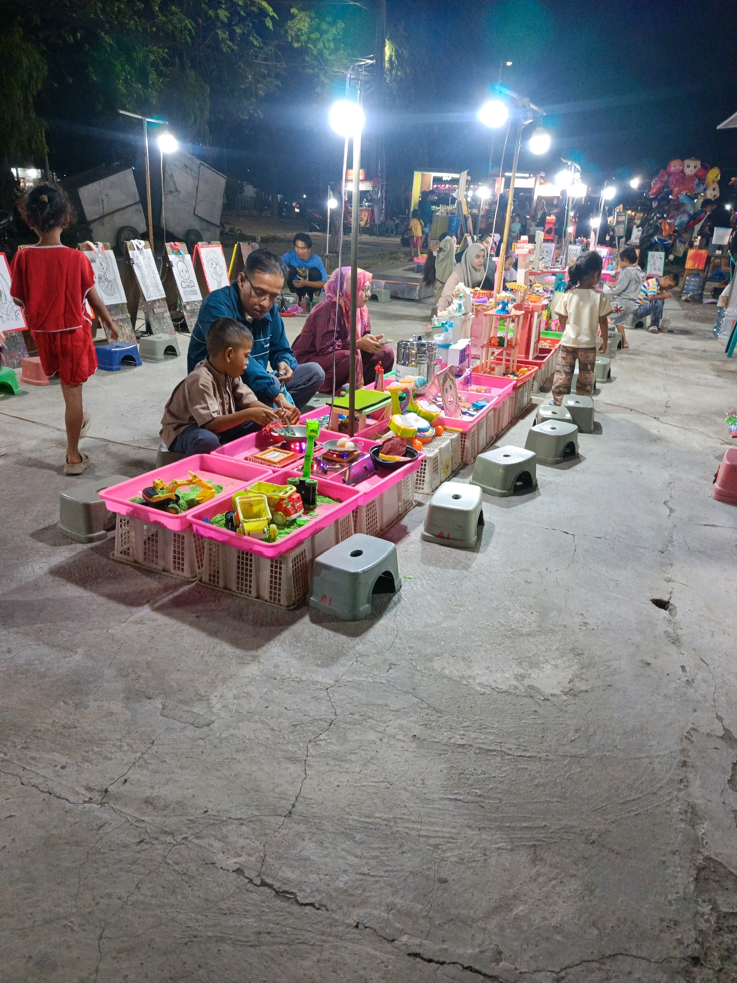 The image depicts a night scene at what appears to be an outdoor market or fair in Sedati Agung, Indonesia. The ground is a worn, cracked concrete surface, illuminated by bright overhead lights, possibly street lamps or vendor lights, casting harsh shadows. In the foreground, a group of people are gathered around a display of toys. A man in a dark blue shirt and glasses is seated, interacting with a young boy wearing a light brown shirt and dark pants. The boy is focused on playing with a toy set laid out in pink plastic bins. These bins contain various colorful toys, including toy construction vehicles and what appears to be kinetic sand.
To the left of the man and boy, a young girl in a red dress stands with her back to the camera, facing a row of easels displaying drawings. Further back, a woman in a pink hijab is seated and also engaged with the toys. To the right of the main group, other individuals are visible, including a woman in a beige hijab and children browsing or playing with toys on a raised platform. In the background, there are various stalls and structures associated with a market, including what looks like food vendors and stands selling balloons. The overall atmosphere suggests a lively, family-oriented event at night.