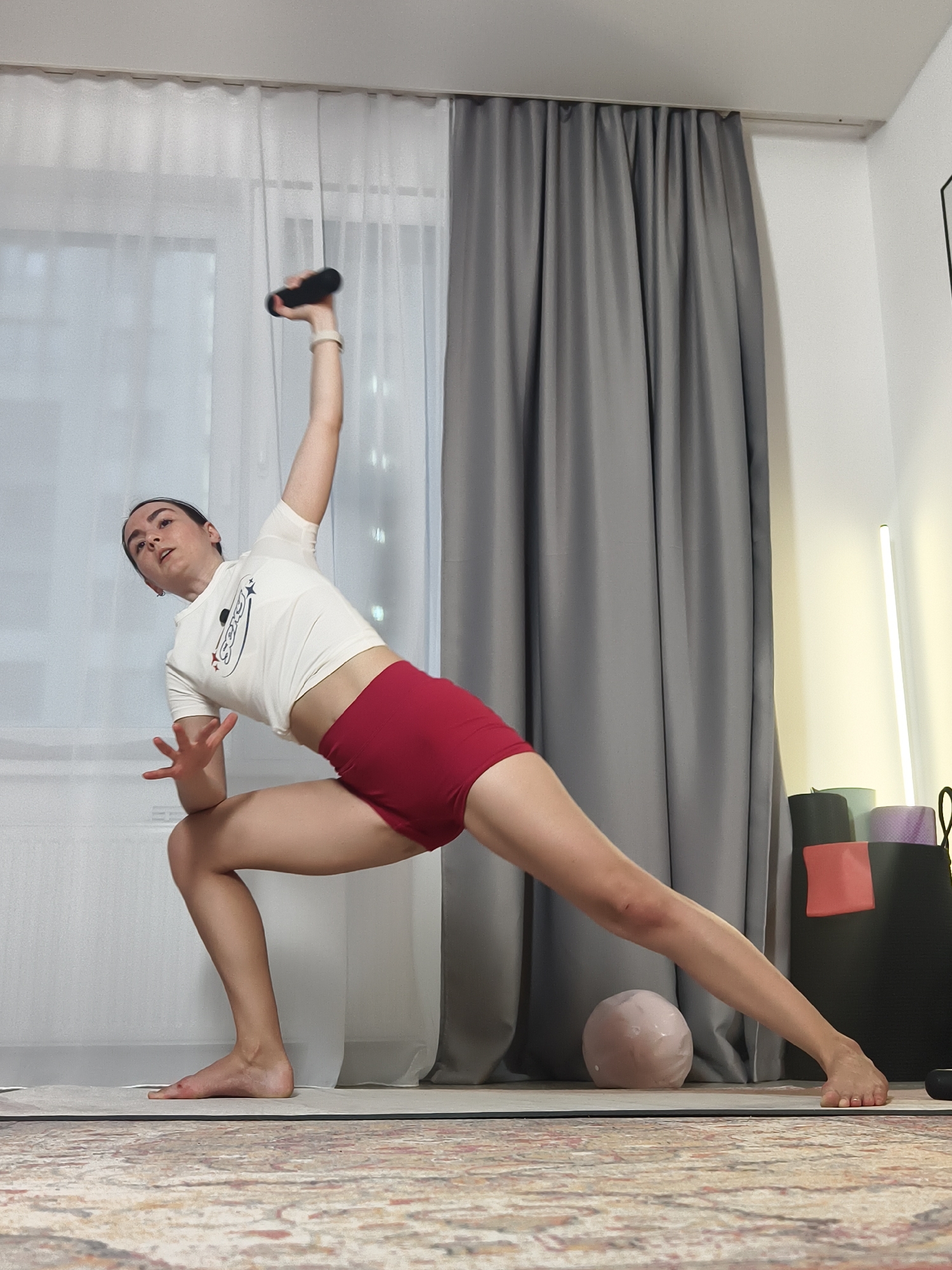 A woman is performing a yoga pose indoors. She is wearing a white cropped t-shirt with a logo, and red athletic shorts. Her left arm is bent at the elbow with her hand near her face, and her right arm is raised above her head holding a black object. Her left leg is bent at the knee, while her right leg is extended straight to the side. She is standing on a yoga mat, which is placed on a patterned carpet. In the background, there is a window with sheer white curtains, as well as gray curtains on the right side. A black bag with rolled-up yoga mats is visible in the background, along with a thin vertical light fixture. A light-colored round object is near her extended leg. Based on available online resources, there is no evidence to confirm the location is in Tyumen, Russia.