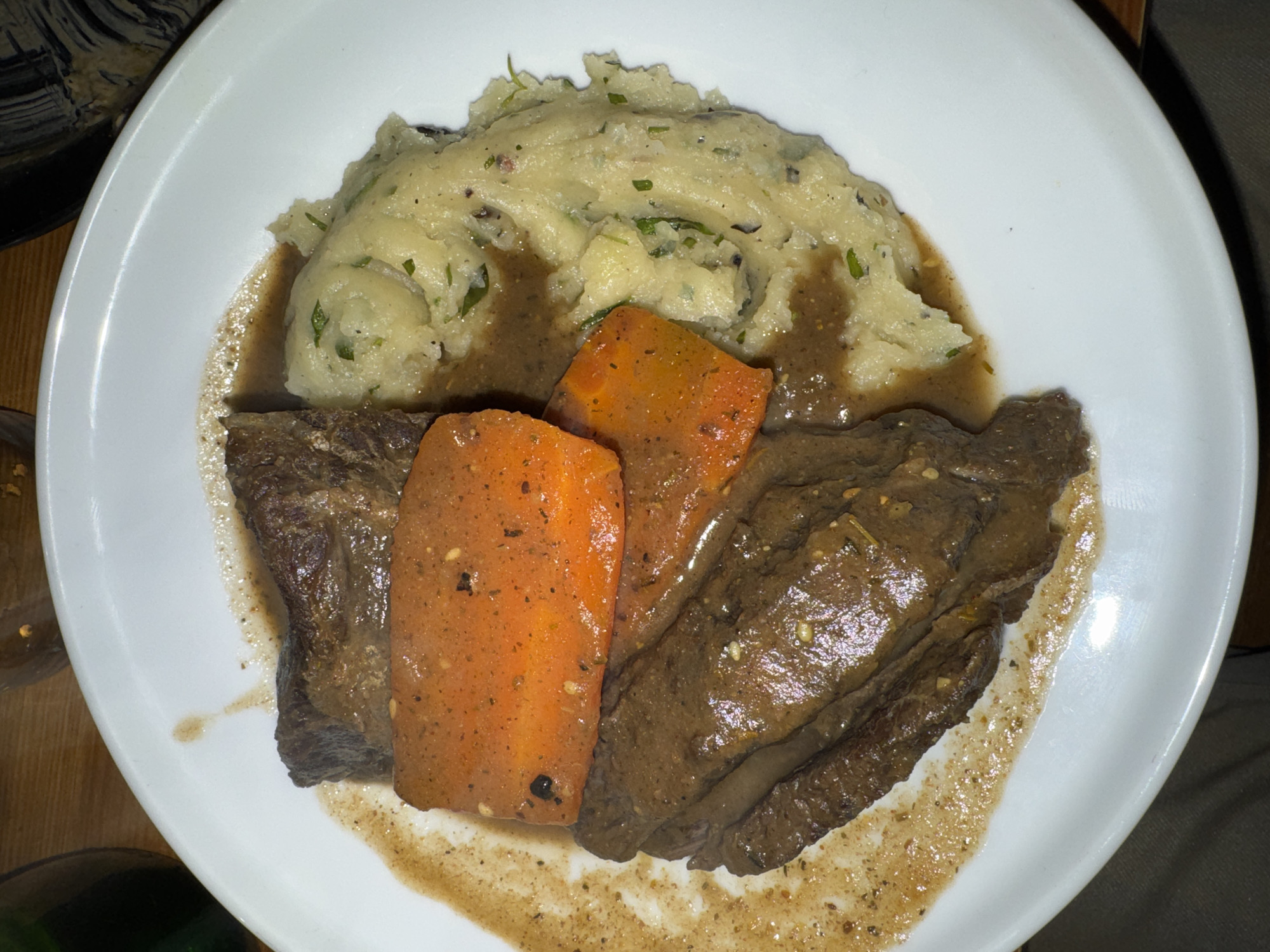 A white oval or rounded rectangular ceramic plate holds a prepared meal. On the left, two pieces of dark brown braised meat are coated in a rich, dark brown gravy exhibiting visible seasoning. Overlapping the meat are two distinct, rectangular cuts of cooked bright orange carrots, similarly glazed with the dark brown gravy and sprinkled with dark spices or herbs. To the right, a substantial portion of light yellowish-white mashed potatoes is present. The potatoes display a textured, rustic consistency and are generously mixed with finely chopped green herbs and small dark specks. The dark brown gravy pools around the meat and carrots, extending slightly onto the potatoes and across the left surface of the white plate. The plate is positioned on a dark, indistinct surface, with a wooden table surface partially visible in the upper background. A dark green bottle or glass is partially visible on the far upper left. The lighting is artificial, suggesting an indoor dining setting. This scene is located in a dining establishment in Paris, France.