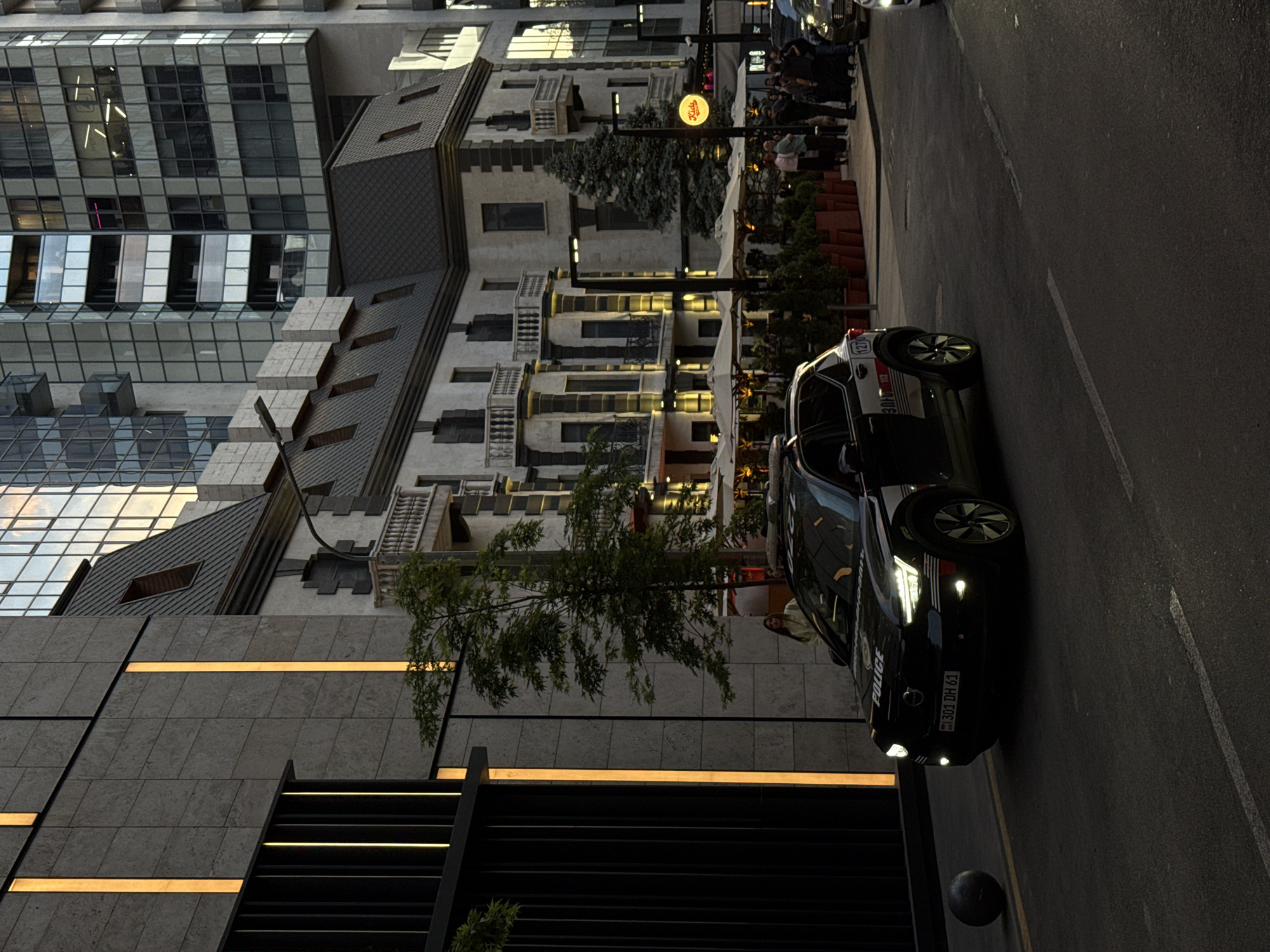 The image depicts a street scene in Yerevan, Armenia. A black police car with license plate "304 DH 61" is parked on the right side of the road. A crowd of people is visible on the sidewalk next to an outdoor seating area with white umbrellas. Buildings with varying architectural styles line the street, including a modern glass-fronted high-rise and a building with a more traditional European design. A "Kids Zone" sign is illuminated above the sidewalk. The overall lighting suggests it is either late afternoon or early evening.