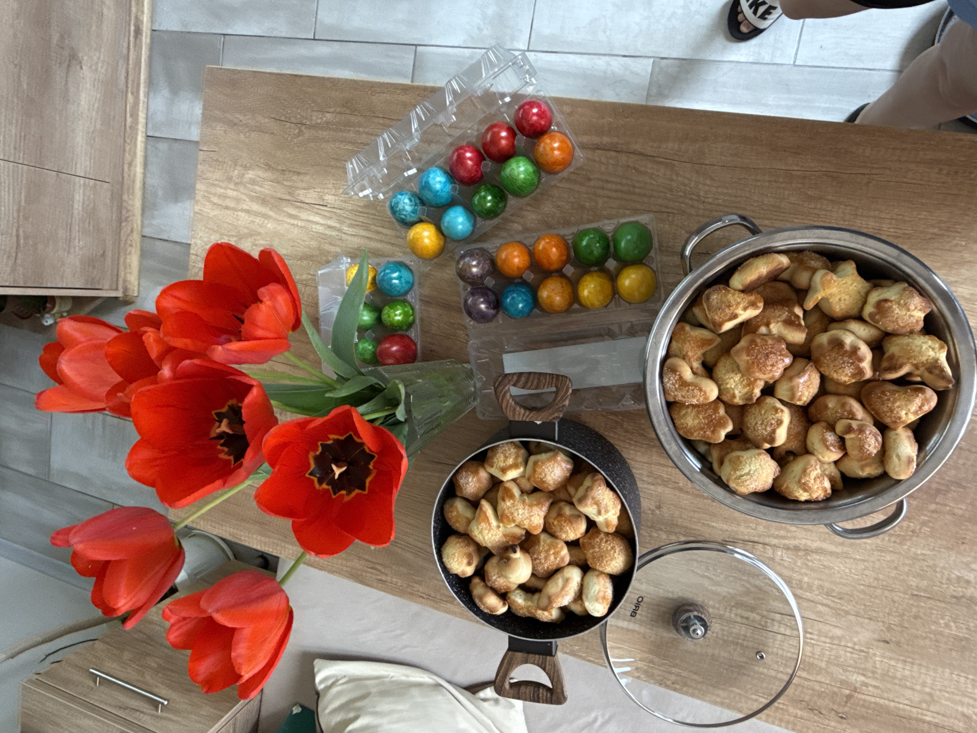 The image shows a wooden table with a vase of red tulips. There are two pots, one black and one silver, filled with baked goods that appear to be small, light-colored cookies. Beside the pots are trays of colorful, spherical objects, possibly painted Easter eggs. The scene is indoors, presumably in a home. Part of a person's legs is visible, wearing sandals. Based on the context of the question (mentioning the city of Stara Zagora, Bulgaria), it can be inferred the image was taken at this location.