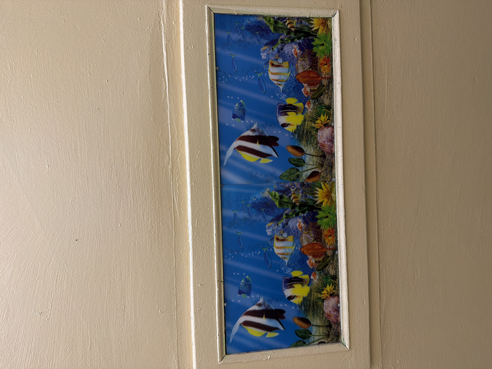 The image shows a close-up view of a beige-painted door or wall panel with a decorative strip featuring an underwater scene with fish. The decorative strip is framed by a thin, light-colored border and appears to be a sticker or decal. The underwater scene depicts various colorful fish, coral, and aquatic plants in a blue, bubbly environment. The painting on the door or wall panel has a textured finish with visible brushstrokes and some minor imperfections, such as small specks. The lighting suggests an indoor setting.