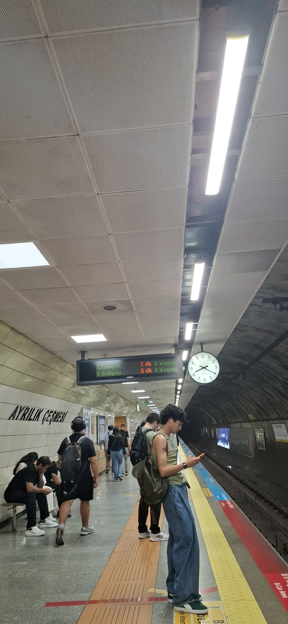 The image depicts a subway platform in Kadıköy, Turkey. Several individuals are present, some seated on a bench and others standing, appearing to wait for a train. One person in the foreground is looking at their phone. A digital display board shows train information. The platform is clean and well-lit, with a clock visible on the wall. The overall atmosphere is that of a typical urban transit station.