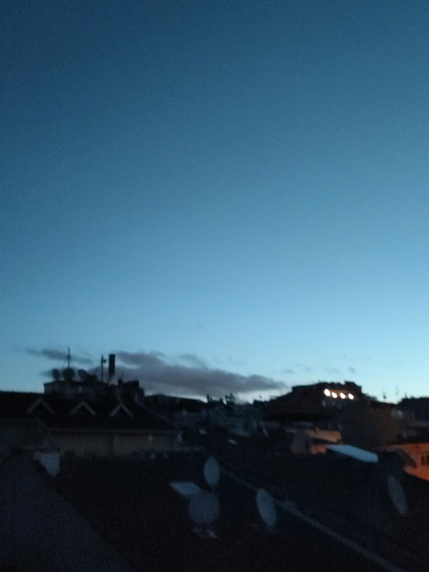 This is a low-light, slightly blurred image of a cityscape at dusk, possibly taken from a rooftop. The sky is a deep blue, transitioning to lighter shades near the horizon. Silhouetted against the sky are buildings with visible roofs, chimneys, and some antenna-like structures. A few illuminated windows or lights are visible on one of the buildings. The foreground is dark and indistinct, showing what appears to be the roof of the vantage point, with some circular or oval shapes that might be reflections or objects. The scene suggests an urban environment, and the presence of satellite dishes on the rooftops might indicate residential or commercial buildings. The overall impression is one of twilight in a populated area.
