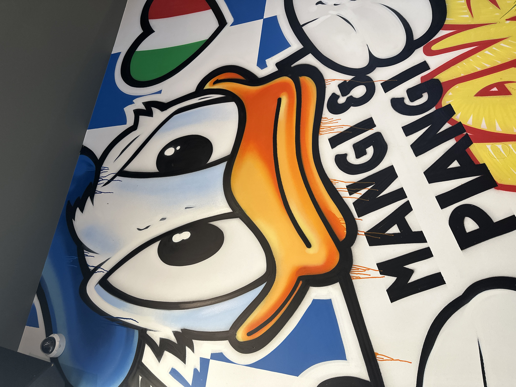 A close-up shot of a mural with cartoon elements. The mural features a stylized duck character with prominent eyes and an orange beak. The character's face appears to be on a blue and white checkered background. Above the duck's head is a symbol with the colors of the Italian flag. To the right of the duck is the word "MANGIA," likely part of a larger phrase. The mural appears to be on a wall next to a light gray wall. The image does not provide enough detail to ascertain its location in Sofia, Bulgaria.