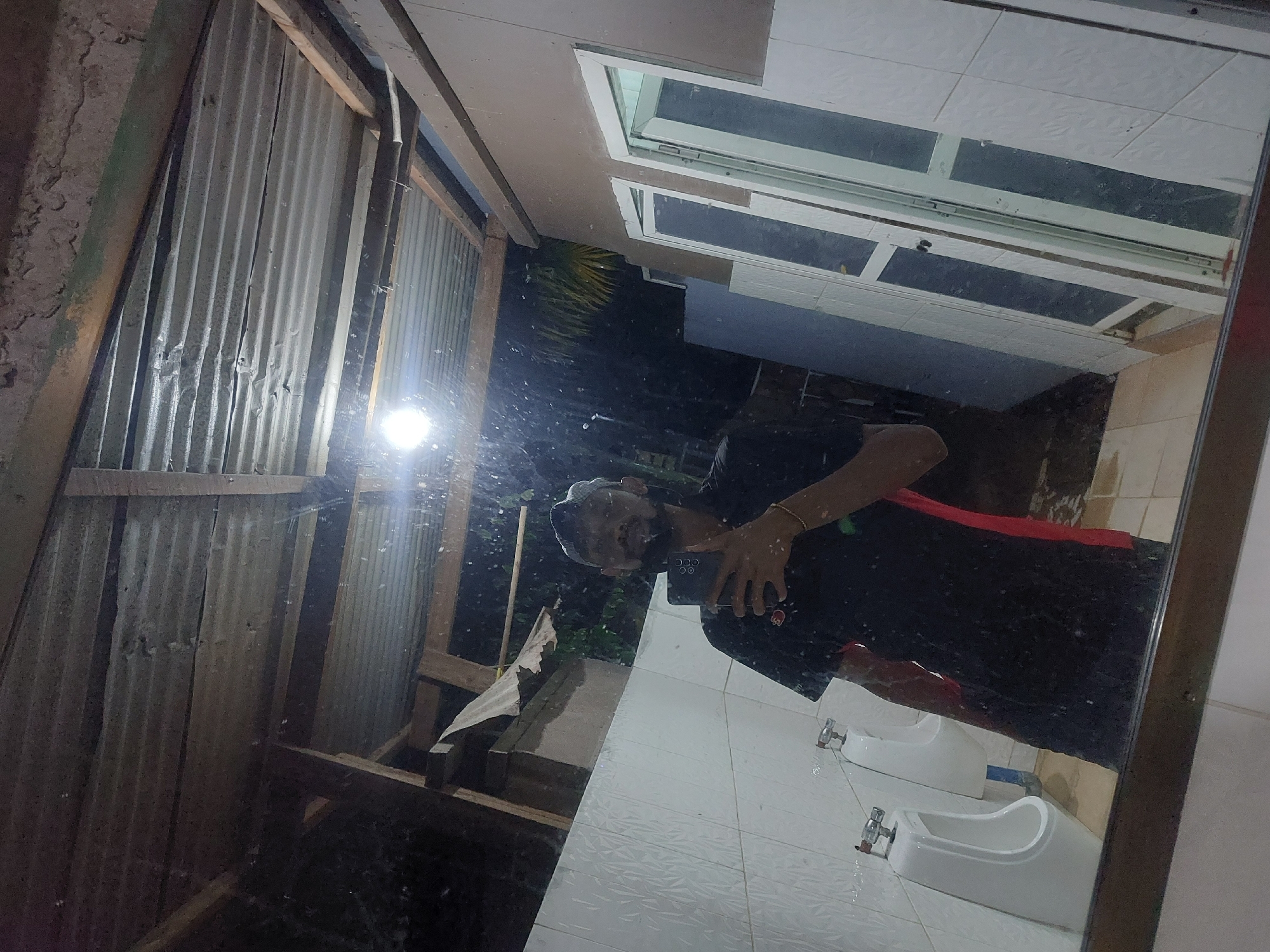 A person is taking a mirror selfie in a bathroom. The bathroom has two white wash basins installed on a white tiled wall. The mirror reflects the exterior of the building which appears to be constructed of corrugated iron. The exterior also shows foliage. The location is Dar es-Salaam, Tanzania.
