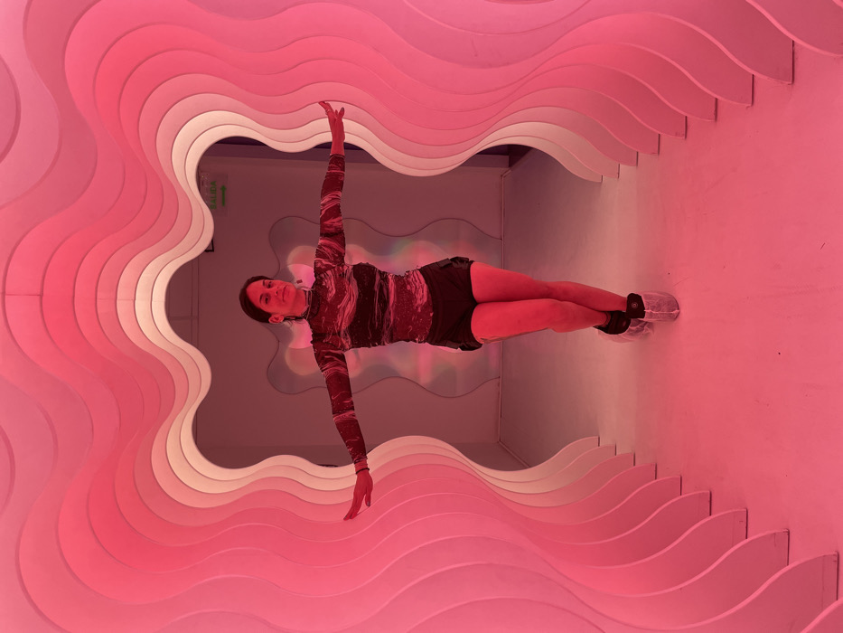 A woman in a marbled long-sleeved shirt and black shorts balances horizontally in a room with a wavy pink tunnel. She extends one arm upward and the other downward. She wears clear plastic shoe covers. The room is lit with pink light, highlighting the wavy tunnel structure that forms the background. Her body aligns with the center of the tunnel, creating a symmetrical composition.
