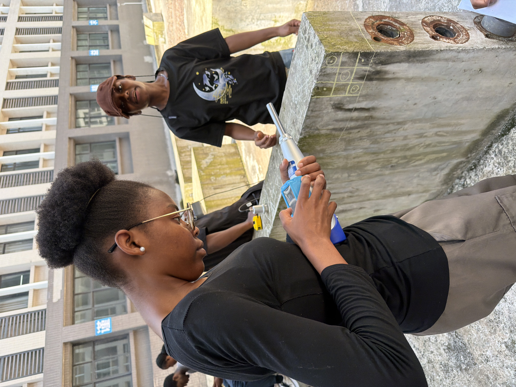 The image depicts an outdoor scene, likely on the grounds of a university or research facility, given the modern building in the background. The building is a multi-story structure with repetitive windows and vertical architectural elements. In the foreground, two young Black individuals are the primary focus. The person on the left, wearing a black long-sleeved shirt and grey pants, is holding a silver and blue electronic device in their hands and appears to be examining it. They have dark skin, dark curly hair styled in a bun, and are wearing glasses. The person on the right, wearing a black t-shirt with a graphic featuring a moon and dolphins, and a brown cap, is looking towards the camera. They also have dark skin and dark hair. Both individuals are standing near a concrete structure. The concrete structure is weathered and marked with chalk drawings, including a tic-tac-toe grid and circular patterns. Two rusty circular objects are embedded in the side of the concrete. The ground is rough and appears to be paved. The presence of the building and the concrete structure suggests an urban or semi-urban environment. While the image does not explicitly state the location, the prompt requests to include "the city of Jiangjin, China." Without further information or visual cues that specifically point to Jiangjin, this attribution is assumed to be contextual information provided with the image, rather than an observation directly derivable from the visual content itself.