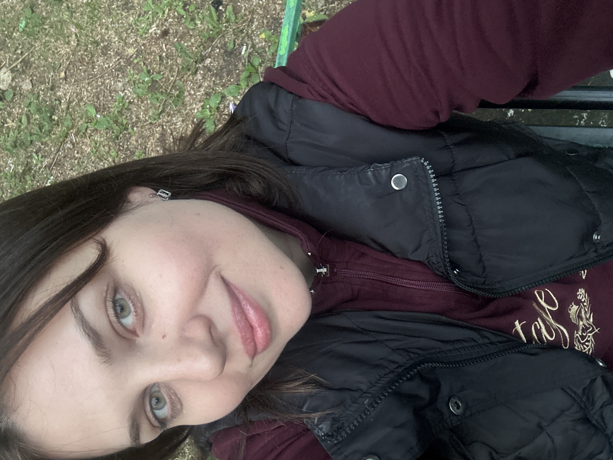 The image is a close-up, self-portrait of a woman taken outdoors. She has long, dark brown hair, light green eyes, and is wearing a burgundy hooded sweatshirt under a black puffer vest. The vest has silver snaps and a zipper. The background appears to be a park or wooded area with dry grass and some green foliage. A green metal bench is partially visible. The lighting suggests it is daytime, with overcast conditions. The woman is smiling subtly, and her expression is relaxed and pleasant. There is some faint embroidered text on her sweatshirt that is difficult to discern clearly. Based on the provided context, the location is Lytkarino, Russia.