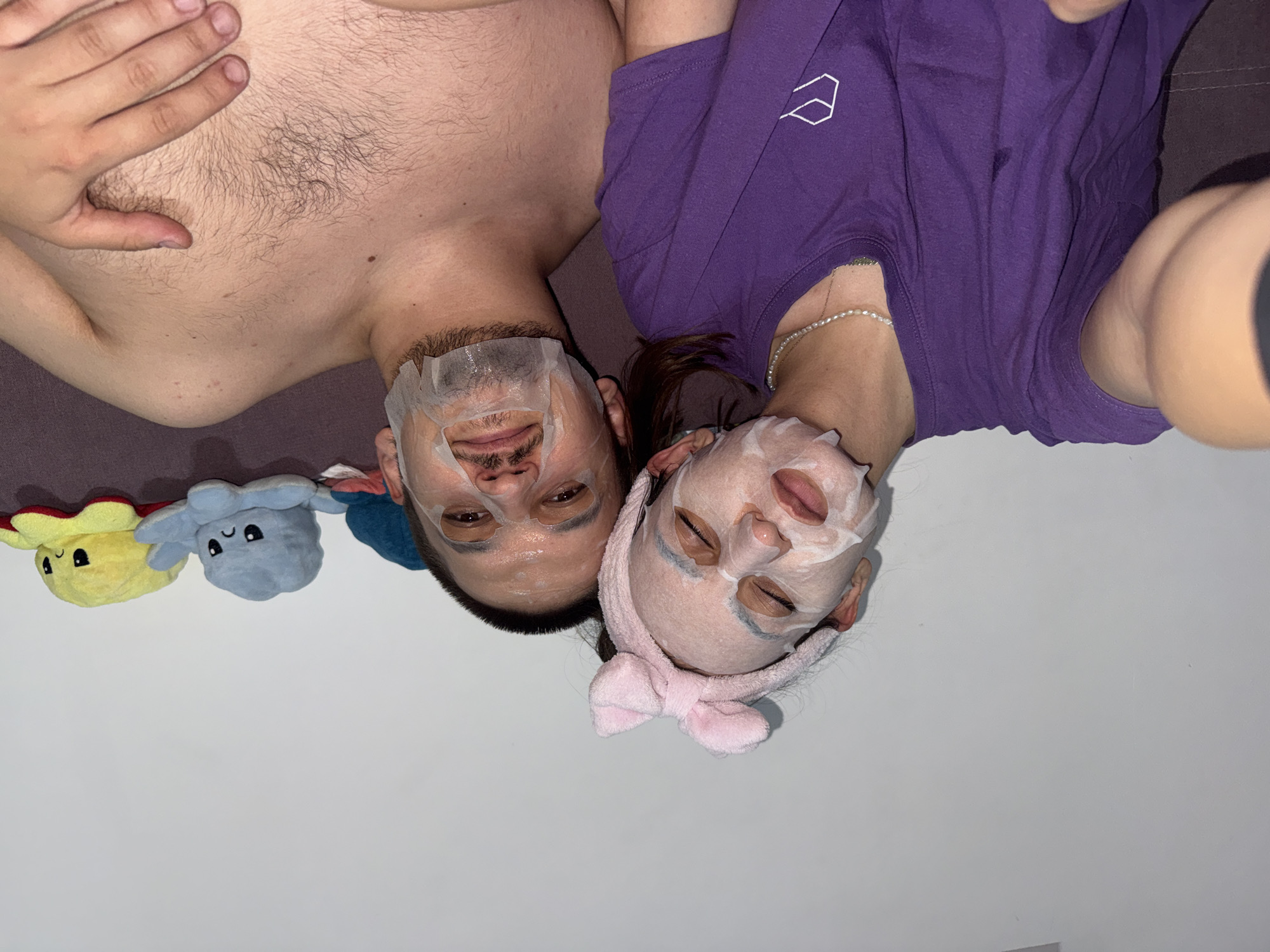Two people, a male and a female, are lying down on a couch, both wearing facial masks. The male is topless with chest hair visible. The female is wearing a purple t-shirt and a pearl necklace. A pink bow headband is on the female's head. Two reversible octopus plushies, yellow and blue, are placed beside them. The background is a light-colored wall. The location is not identifiable as Plovdiv, Bulgaria based on the visual information provided.