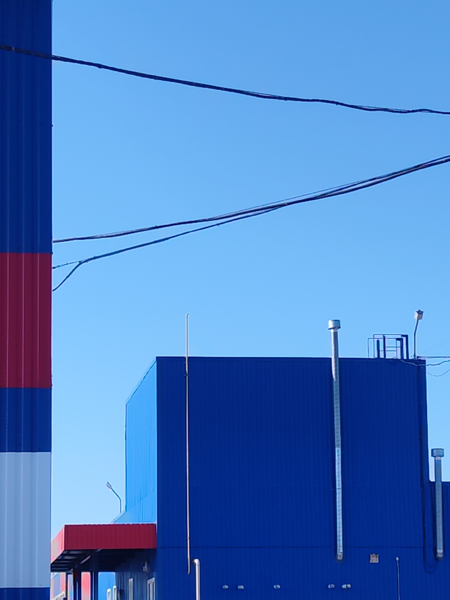 The image depicts a view of a building and a tall chimney structure in Chaikovsky, Russia. The building is predominantly blue with vertical corrugated panels, featuring a red awning and some ventilation pipes on the exterior. A small platform with railing and a light fixture are visible on the roof. The chimney, located on the left of the frame, has distinct bands of blue, red, and white. Overhead, a couple of power lines stretch across the clear, blue sky. No people are immediately visible.