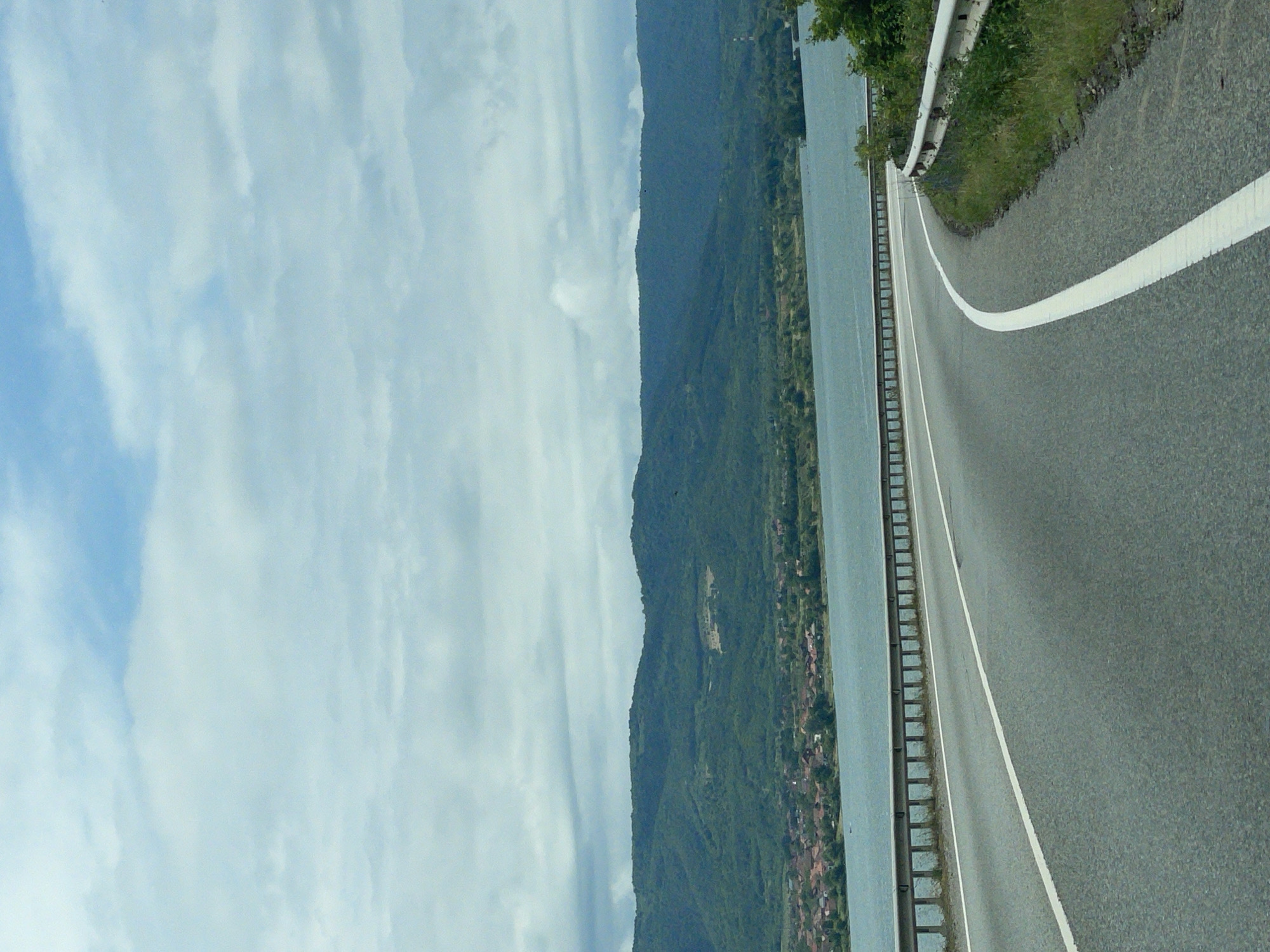 The image captures a panoramic view of a vast body of water, likely a lake or river, bordered by a large mountain range under a partly cloudy sky. The sky is a mix of light blue and white, with diffused clouds. The mountains are covered in dense green vegetation, indicating a lush landscape.
On the right side of the frame, an asphalt road curves gently, following the contours of the shoreline. A solid white line delineates the edge of the road closest to the water. A metal guardrail runs along the side of the road, providing a barrier between the roadway and the water.
Along the base of the mountains, adjacent to the water, several small settlements or villages can be seen, characterized by clusters of buildings with reddish-brown roofs. The scene appears to be set during daytime, with clear visibility and natural light. There are no people visible, and no specific activities are taking place; it is a static landscape shot, possibly taken from a moving vehicle. No text is visible in the image. The location context indicates this is a view from Drobeta-Turnu Severin, Romania, suggesting the body of water is the Danube River.