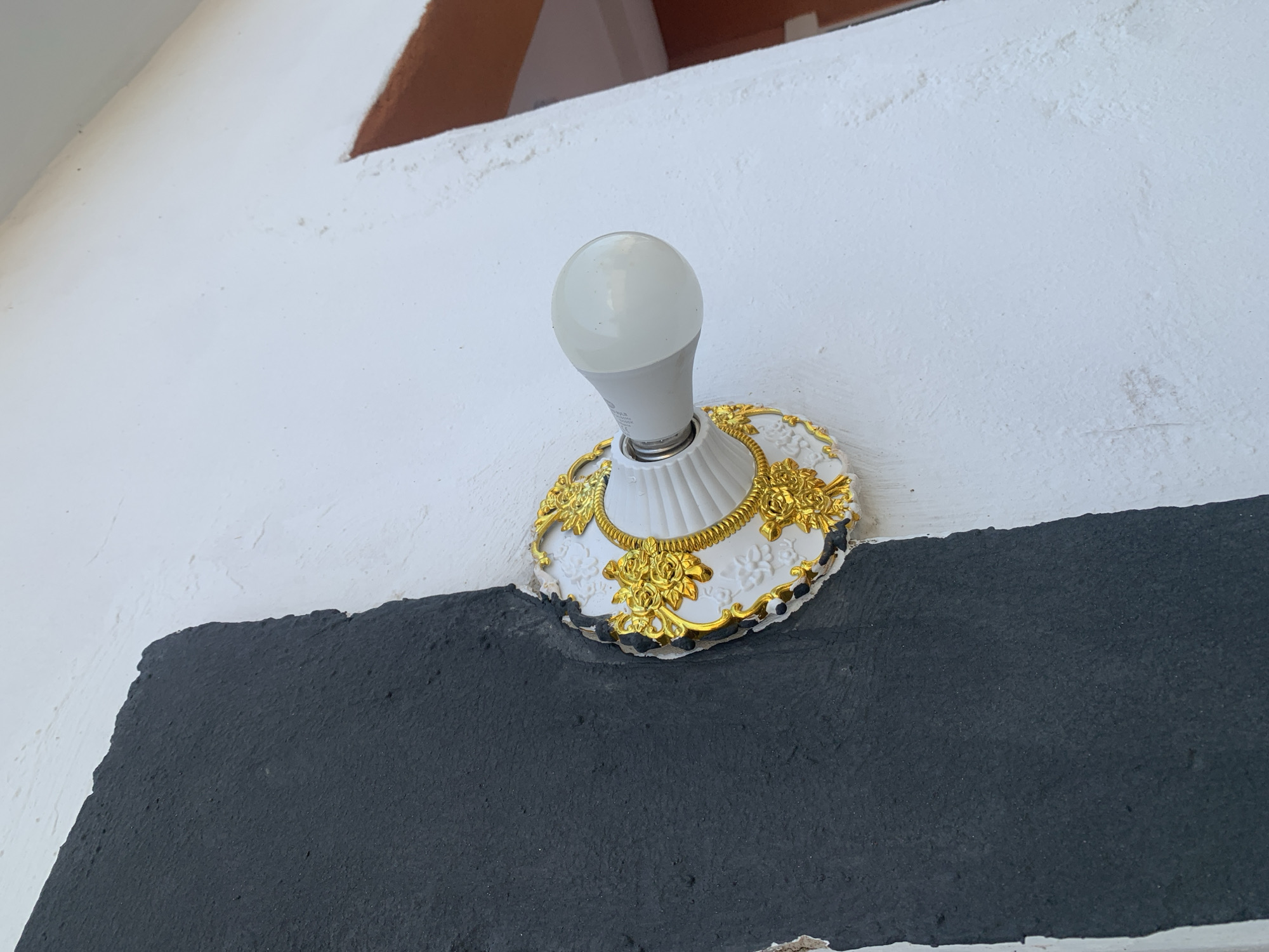 An outdoor close-up shot shows a white and gold decorative fixture with a white LED lightbulb protruding from the center, mounted on a textured white wall. To the left of the fixture, a section of the wall is painted a rough, textured black, with some of the black paint chipping away at the edge where it meets the white wall and the decorative fixture. The black paint appears to be a thicker, more uneven application compared to the white paint. The fixture itself is ornate, with raised floral patterns in a shiny gold finish surrounding the white base. The lightbulb is simple and cylindrical with a rounded tip. In the upper right corner of the image, a sliver of a reddish-brown frame or architectural detail is visible against the white wall. The lighting suggests it is daytime, casting soft shadows.