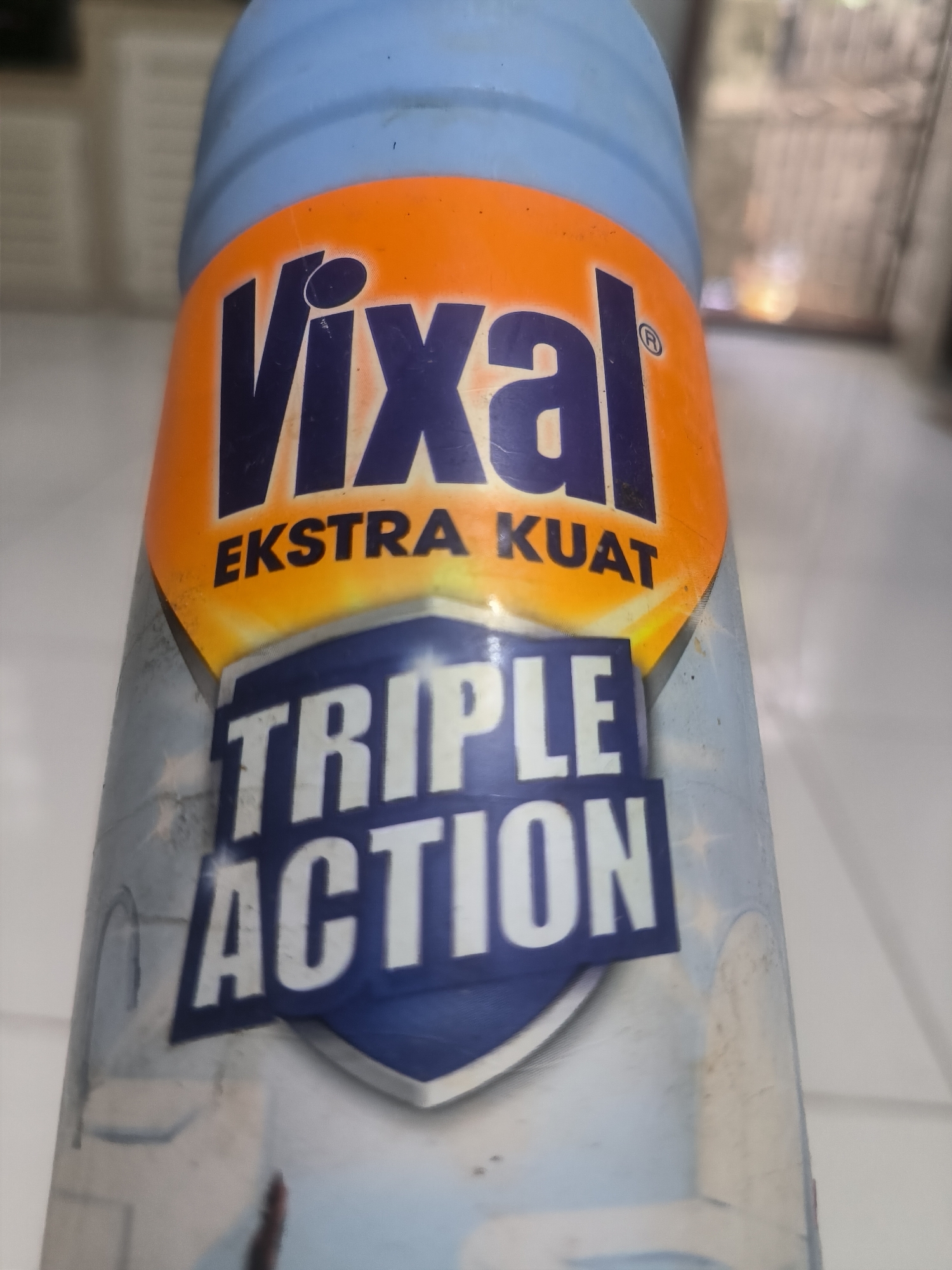 The image shows a bottle of Vixal toilet bowl cleaner. The label reads "Vixal EKSTRA KUAT" above a shield design that contains the words "TRIPLE ACTION". The bottle is light blue at the top and transitions to a white background below the label. The background is blurry and indistinct, but appears to be a tiled surface, possibly indoors. The location of the city of Lhokseumawe, Indonesia, is not evident from the image.
