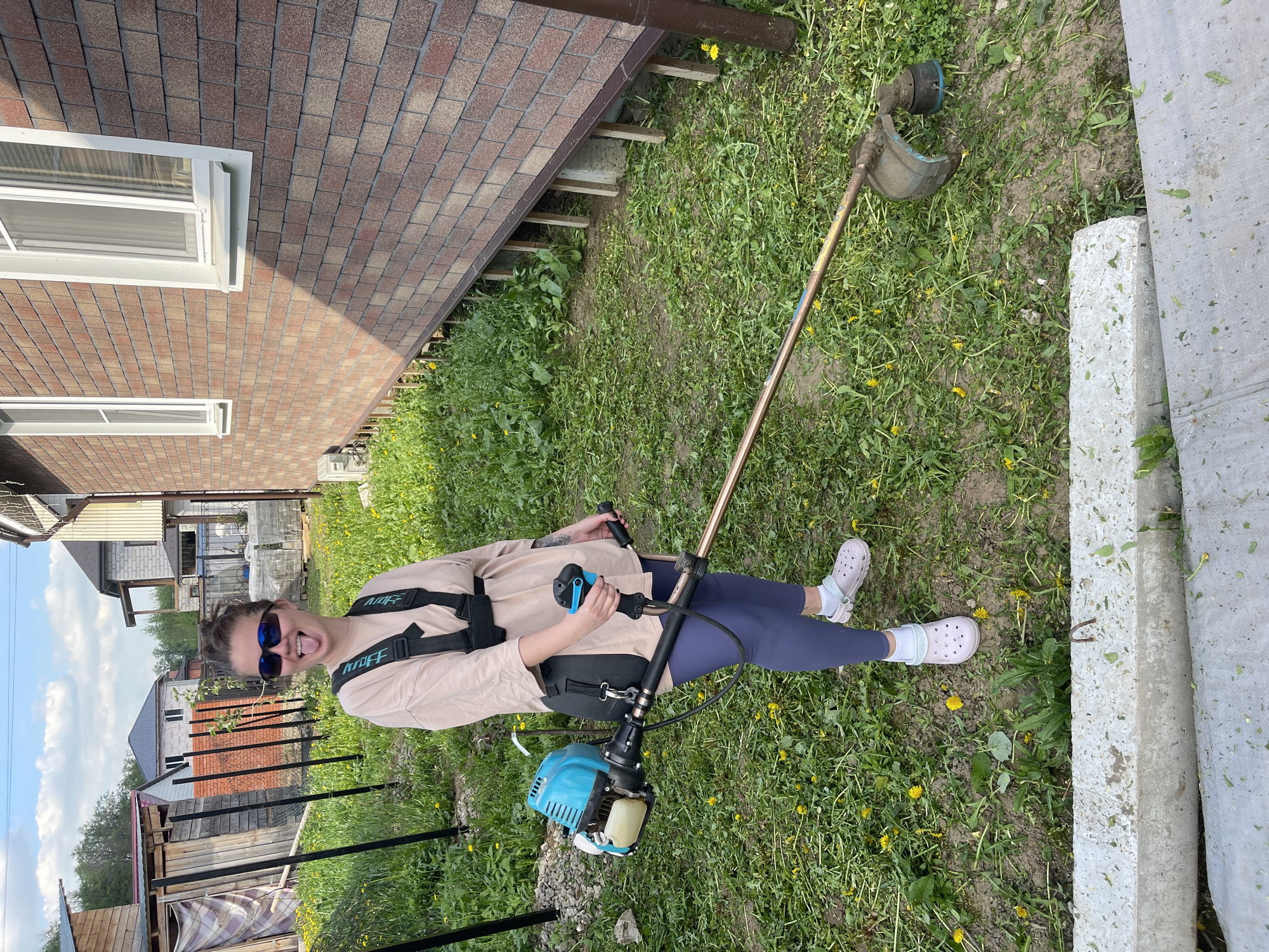 A woman stands in an overgrown yard in the city of Мосметаллоконструкция Онт, Russia, operating a string trimmer. She wears sunglasses, a light brown long-sleeved shirt, navy leggings, and white clogs with white socks. A harness supports the blue and gray string trimmer. The surrounding environment includes a brick building, grass with yellow dandelions, concrete blocks, and fences suggesting residential properties.