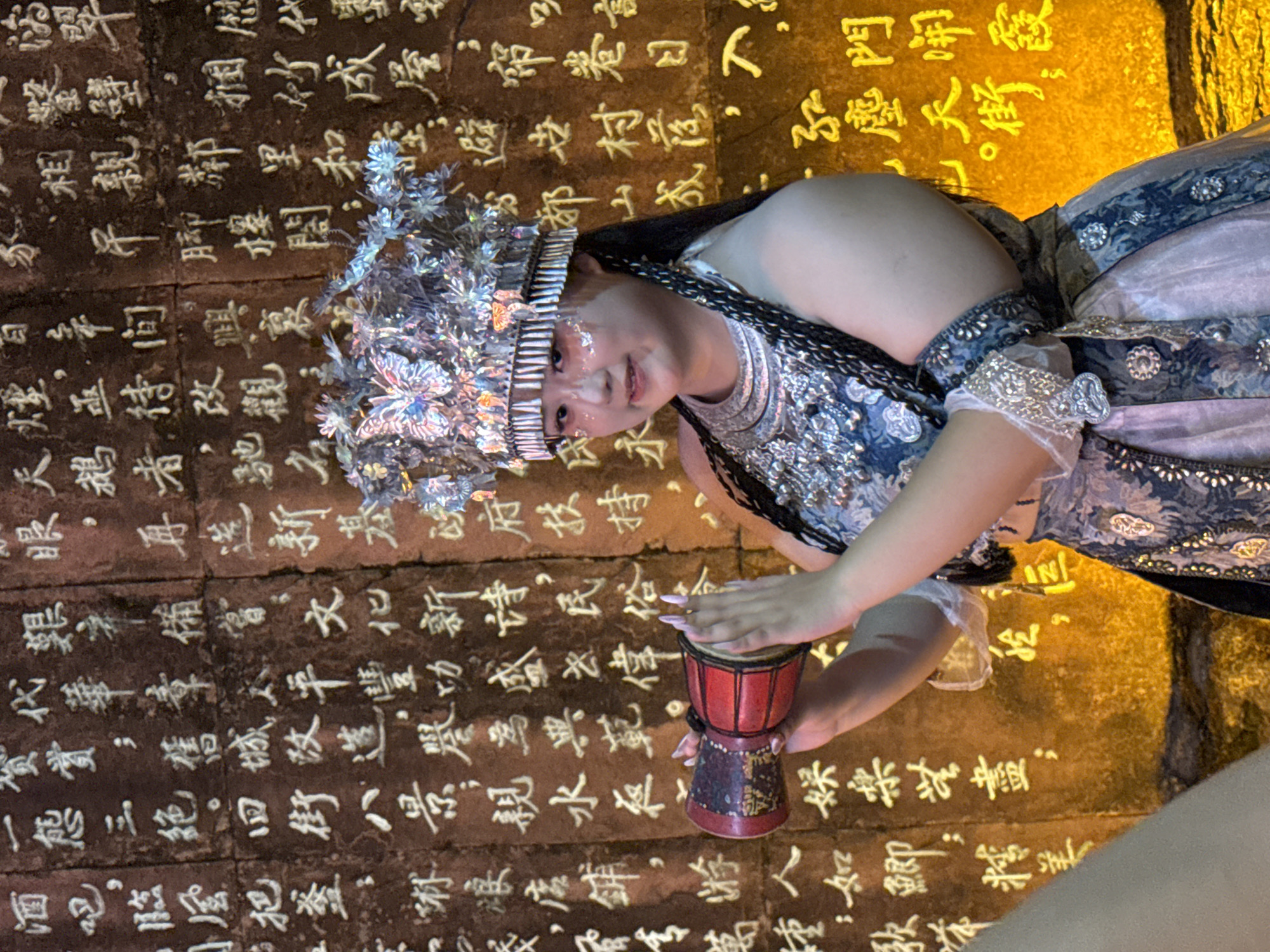 A young woman in traditional Chinese attire, including an ornate silver headdress adorned with butterflies, stands before a wall covered in large, white Chinese characters. She is holding a small red drum with a black and gold band in her hands, her fingers poised as if about to play it. Her face is lightly made up with glitter on her cheeks. She is wearing a blue and silver patterned garment with decorative trim. The wall behind her appears to be made of stone or plaster, with the characters carved or painted onto its surface. The overall lighting is warm and yellowish, suggesting an indoor or nighttime setting. The characters are densely packed, creating a textured backdrop. The image seems to capture a moment during a cultural performance or ceremony. The location is identified as Yuzhong District, China.