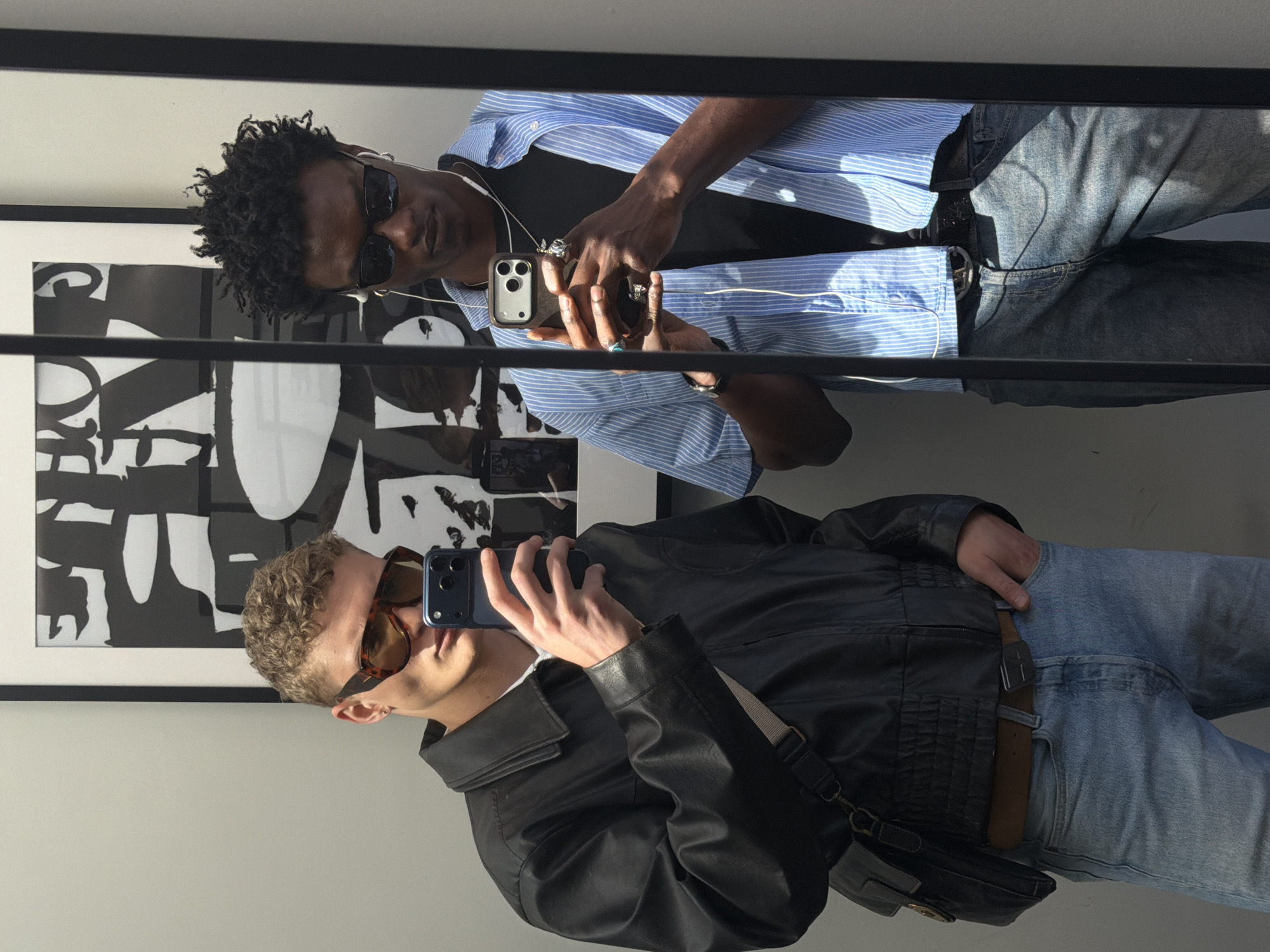 Two men are captured in a mirror selfie, standing side-by-side in what appears to be an indoor setting in London, United Kingdom. The image is taken from a slightly rotated perspective, showing the subjects angled.
The man on the left has dark skin and short, curly dark hair. He wears black sunglasses, a light blue and white striped long-sleeved shirt unbuttoned over a black t-shirt, and light blue jeans. White wired earphones are visible around his neck. His hands are holding a silver-backed smartphone, and he wears multiple rings on his fingers.
The man on the right has fair skin and short, curly light brown hair. He wears brown-tinted, tortoise-shell framed sunglasses and a black leather jacket over a white shirt. He is also dressed in light blue jeans and has a black shoulder bag with a brown strap across his body. He holds a dark blue smartphone, seemingly taking his own mirror selfie.
Both men are looking towards their respective phones, presumably checking their poses or the photo. Behind them, a large framed abstract artwork with bold black and white shapes is visible on a white wall. The scene is brightly lit, likely by natural light, casting distinct shadows and highlighting details of their clothing and accessories. No discernible text is visible in the image.