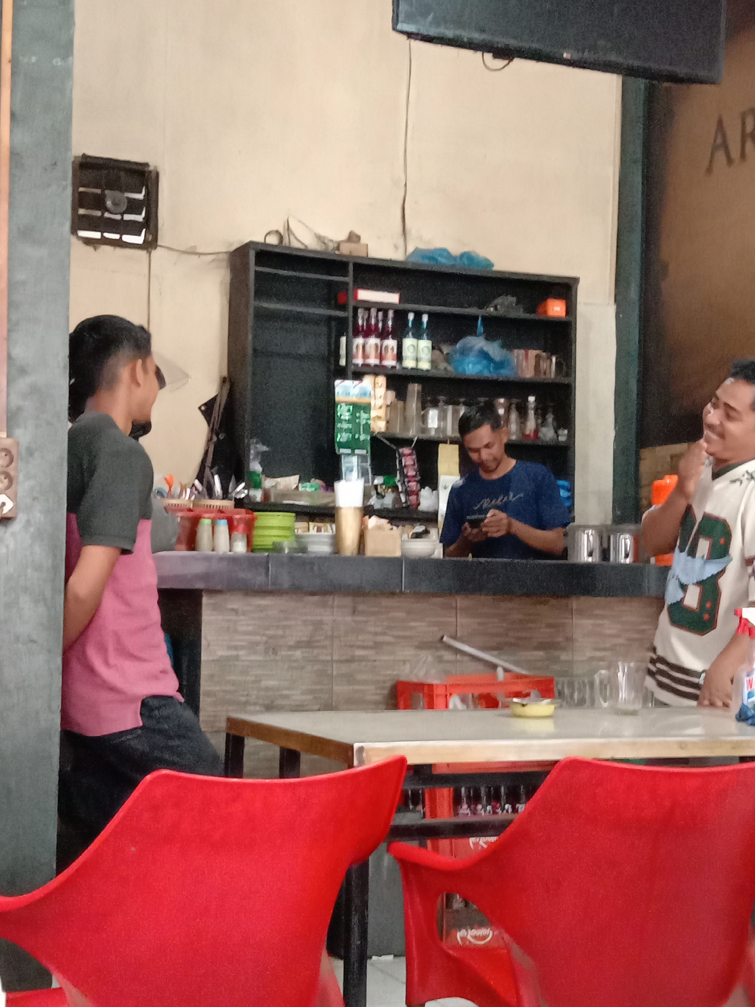 The image depicts the interior of a casual eatery or cafe in Lhokseumawe, Indonesia. In the foreground, two red plastic chairs are visible, one prominently in the lower left and another partially visible in the lower right. Behind the chairs, a counter area is set up with various items. A young man wearing a dark gray t-shirt with a pink t-shirt underneath and dark pants stands to the left, facing away from the camera and looking towards the counter. Behind the counter, a man in a blue t-shirt is looking at his phone. Further to the right, another man, wearing a white t-shirt with green and black accents, is standing and interacting with someone outside the frame. The counter area has shelves stocked with bottles and containers. In the background, there is a large black speaker mounted on the wall. The overall environment suggests a relaxed, everyday setting.