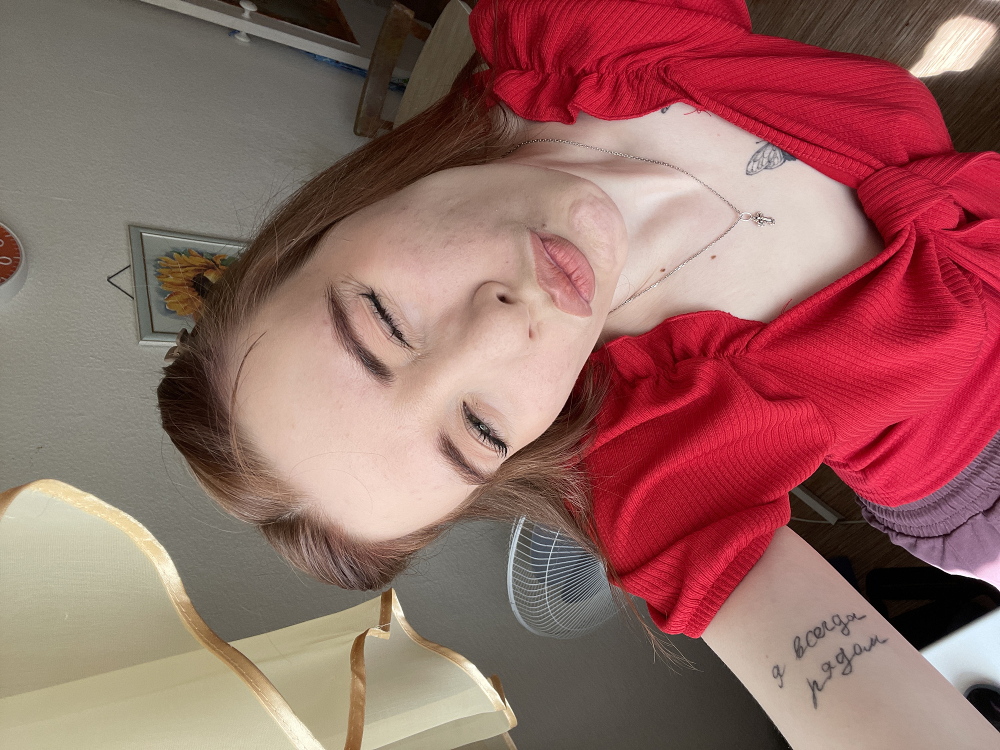 A medium close-up, portrait-oriented shot features a young person with light skin and long, light brown hair, lying down or posing with their head tilted to the right. The person is wearing a bright red, ribbed top with ruffled details around the neckline or sleeves. They have their eyes closed, and their lips are slightly parted and pursed, giving a contemplative or soft expression.
Notable features include visible texture differences and what appear to be scars on the left side of the person's face, particularly around the mouth and cheek area. A delicate silver-colored necklace with a small, indistinct pendant rests on their collarbone, beneath which a dark, butterfly-like tattoo is partially visible on their upper chest. Another tattoo is prominent on their inner left forearm, reading "я всегда рядом" in Cyrillic script, which translates to "I am always close" or "I am always there."
The background suggests an indoor setting, likely a room with light-colored walls. Elements visible include a round, orange clock with white numerals on the wall, a framed picture depicting a sunflower, a white electric fan, and a lamp with a light-colored shade. Natural light illuminates the scene, creating soft shadows, suggesting it is daytime. The floor appears to be made of light-colored wood.