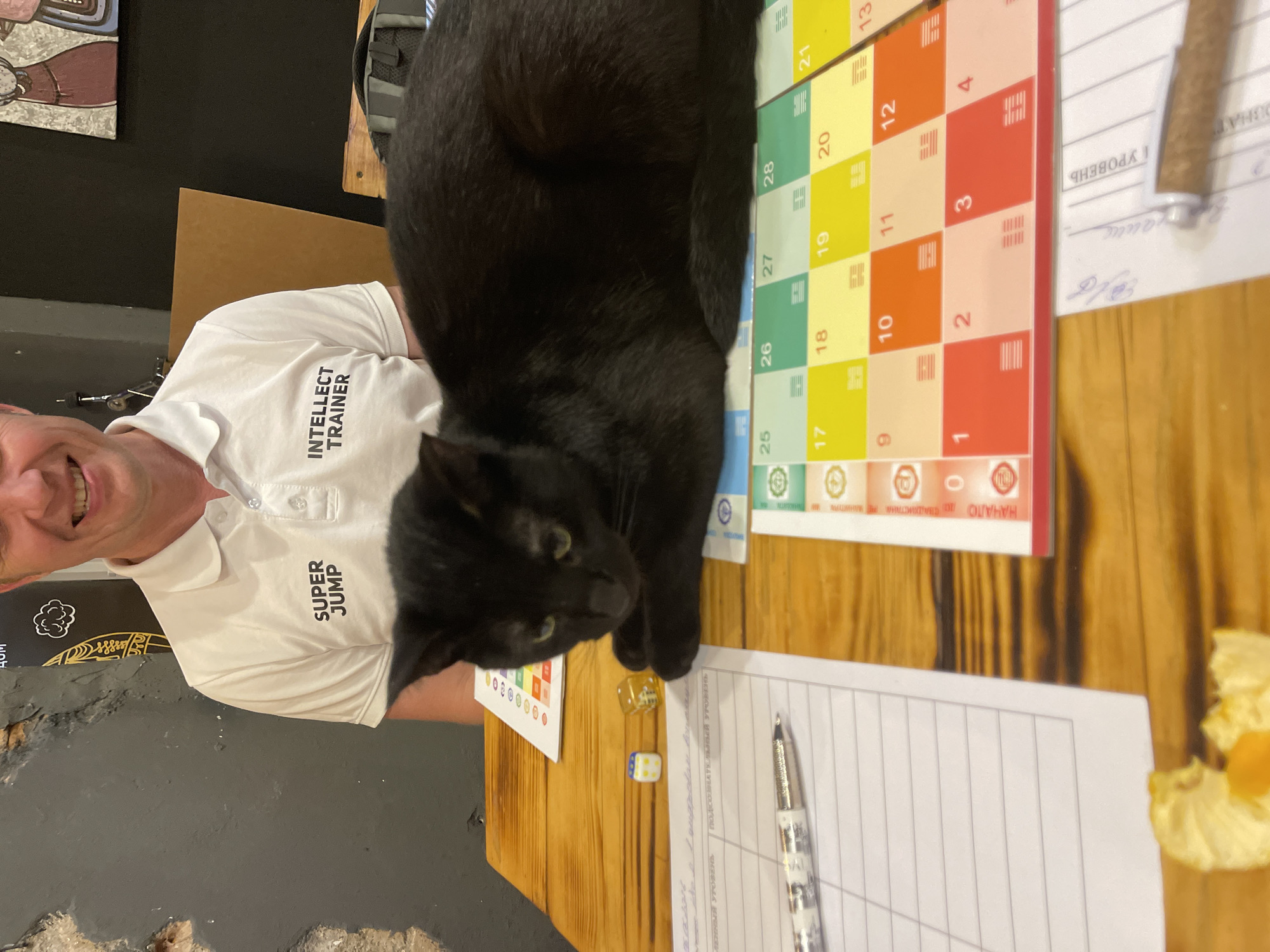 A black cat rests on a board game on a wooden table. A smiling male, partially visible from the nose down, sits behind the table. He wears a white polo shirt with "SUPER JUMP" on the left chest and "INTELLECT TRAINER" on the right. The black cat, with green eyes, lies in a loaf position on a colorful board game featuring numbered squares (1-4, 9-12, 17-21, 25-28 visible), I Ching trigrams, and Russian text including "НАЧАЛО" (START) and "0". On the table, to the left of the cat, are two dice (one white with yellow dots showing '2', one gold with white dots showing '3'), a silver-colored pen, and lined paper with Russian text, including "УРОВЕНЬ" (LEVEL) and "ПОДСОЗНАТЕЛЬНЫЙ УРОВЕНЬ" (SUBCONSCIOUS LEVEL). A peeled citrus fruit segment is visible in the bottom left corner. The background features a dark, textured wall and a painting depicting stylized figures. The scene is set indoors in the city of Kaliningrad, Russia.