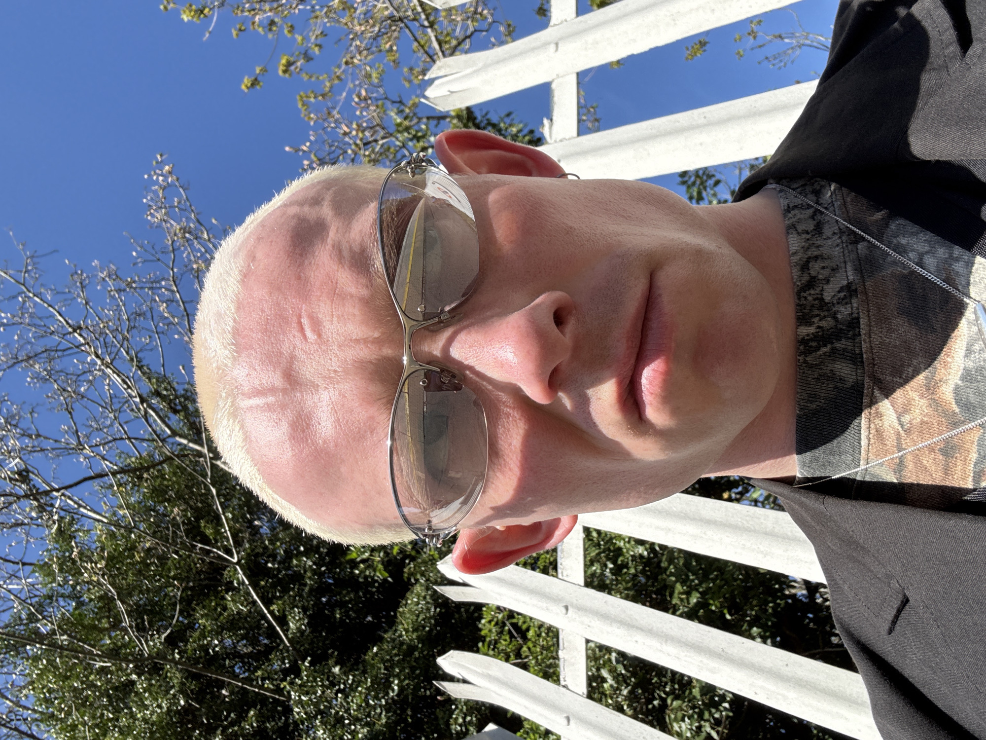 A low-angle, eye-level shot shows a man with a shaved head wearing silver sunglasses and a dark-colored jacket with a camouflage collar. He is standing in front of a white picket fence. Behind the fence, there are trees and a bright blue sky. The image lacks sufficient information to determine the location as the city of London, United Kingdom.