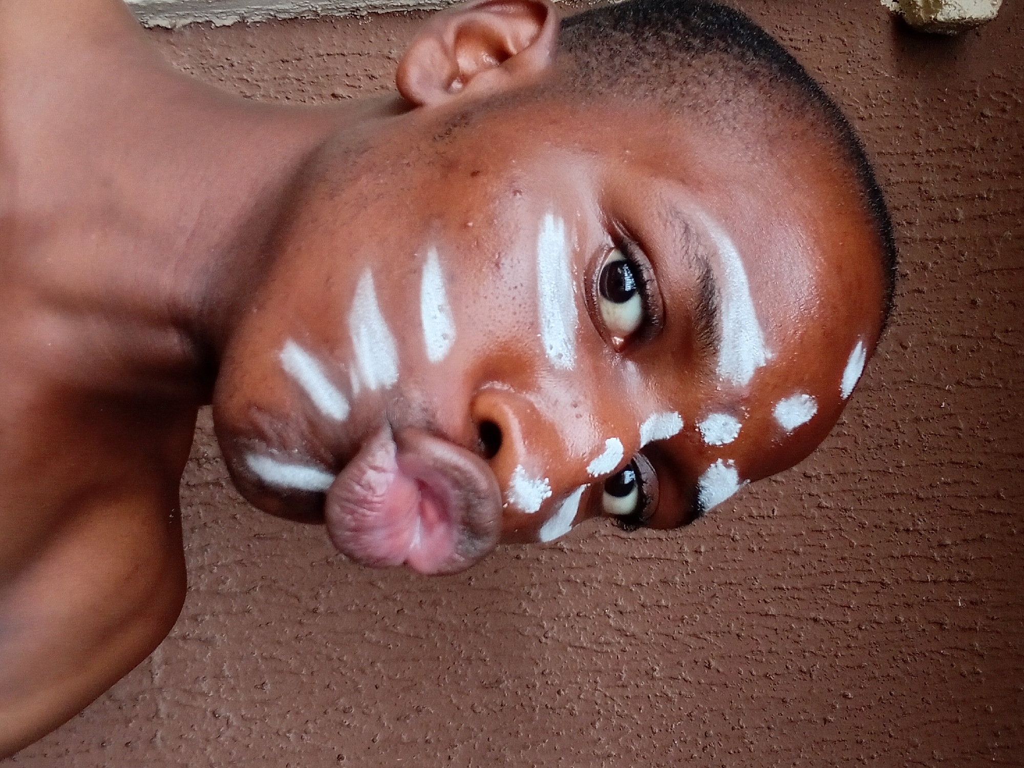 The media file features a close-up, rotated view of a young Black person with white markings on their face. The subject is looking towards the camera with an open mouth, pouting their lips. White paint is applied in a pattern of dots and streaks across the forehead, cheeks, and around the eyes. The skin appears to be slightly glistening, possibly due to sweat or oil. The subject's ear is visible on the left side of the frame, and their neck and shoulder are also partially visible, showing smooth, dark skin.
The background consists of a textured, reddish-brown surface, consistent with a wall or ground, which appears to be outdoors. The lighting suggests it is daytime, as the scene is well-lit. The overall impression is a posed portrait, possibly part of a cultural or artistic expression, given the face paint. No text is visible in the image. The location context is Nkpor, Nigeria.