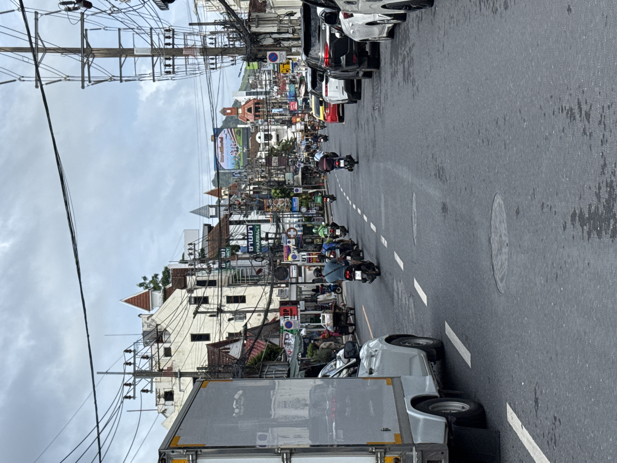 The image depicts a busy street scene in Patong, Thailand, likely on a cloudy day. The foreground is dominated by a white delivery truck on the left and the asphalt road with white lane markings and a circular manhole cover. Multiple motorcycles and a few cars are visible on the road, with riders and drivers engaged in transit. Buildings line both sides of the street, displaying a variety of architectural styles, including some with pitched roofs and dormer windows. Signs in what appears to be Thai and English are present on the buildings, indicating businesses such as pharmacies and restaurants. Overhead, a dense network of electrical wires and utility poles crisscrosses the sky. In the distance, hills are visible under the overcast sky. The overall impression is of a vibrant, bustling urban environment with a mix of commercial activity and daily life.