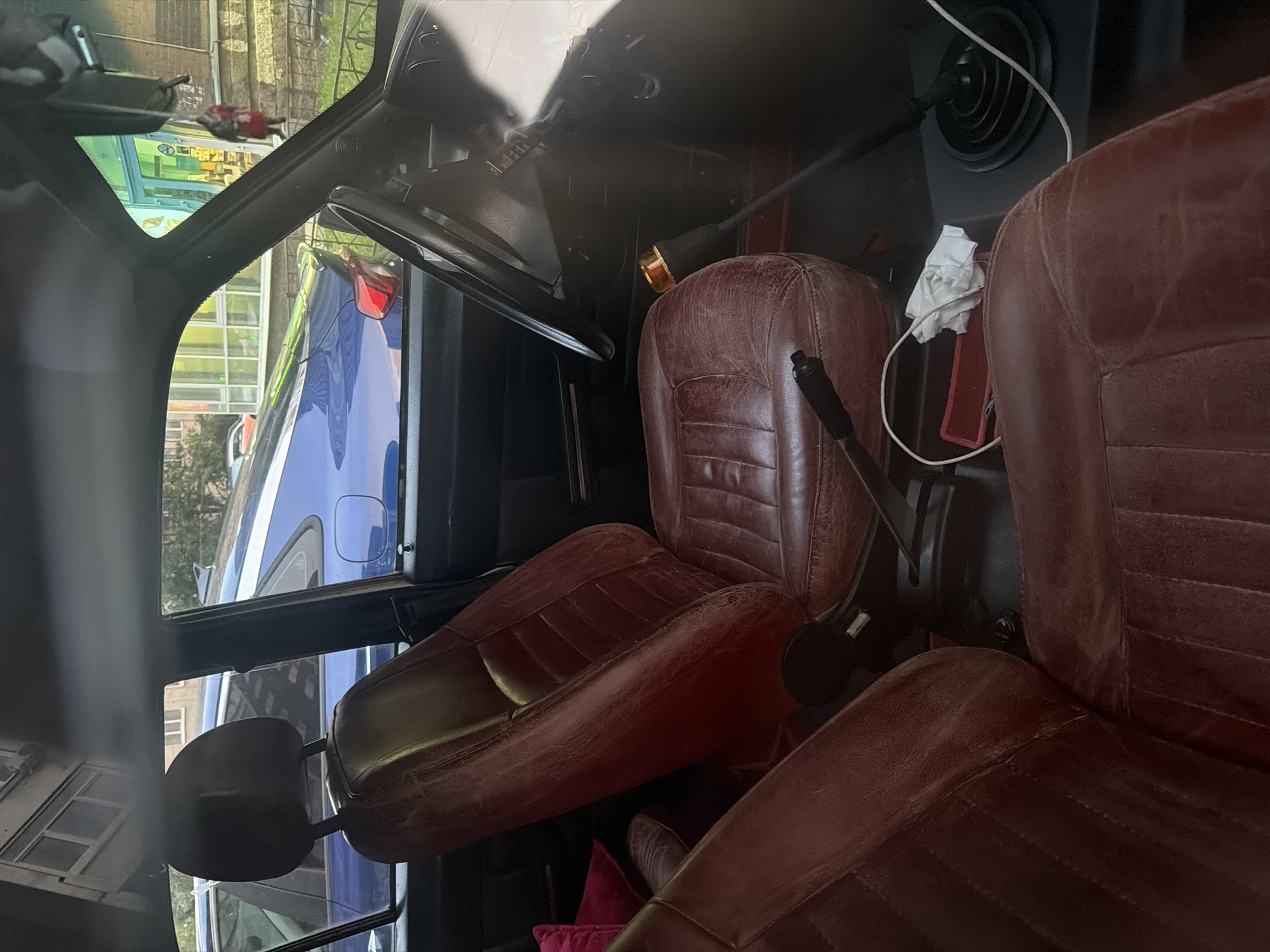 The image shows the interior of a car, specifically the front passenger and driver's seats. The seats are upholstered in dark red leather with a quilted pattern. The dashboard and steering wheel are visible, as is the gear shift. Outside the car, through the windows, a blue car, a building, and trees can be seen. The scene appears to be outdoors, likely in a city setting, given the presence of buildings and other vehicles. The lighting suggests it is daytime. There are no people clearly visible within the car.