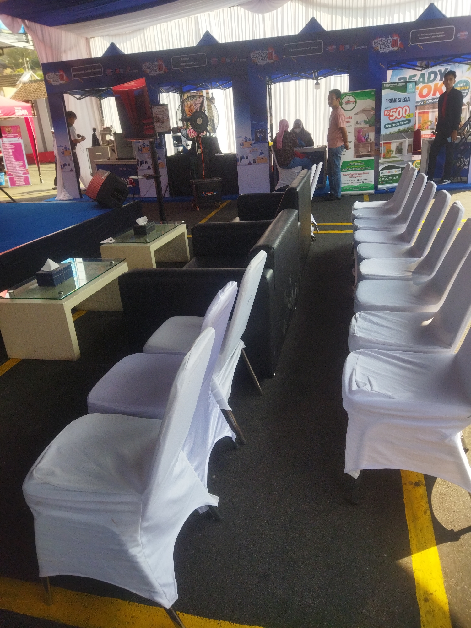 The image depicts an outdoor event setup under a tent in Giripurwo, Indonesia. The scene features rows of white-covered chairs, suggesting a waiting area or seating for an audience. In the background, a blue tent structure with promotional banners is visible, indicating a commercial event or exhibition. Several people are present, including individuals standing near the promotional tent and others possibly involved in setting up or managing the event. There are also tables, a fan, and a speaker, which are typical elements found at such gatherings. The ground is asphalt with yellow markings, characteristic of outdoor event spaces or parking areas.