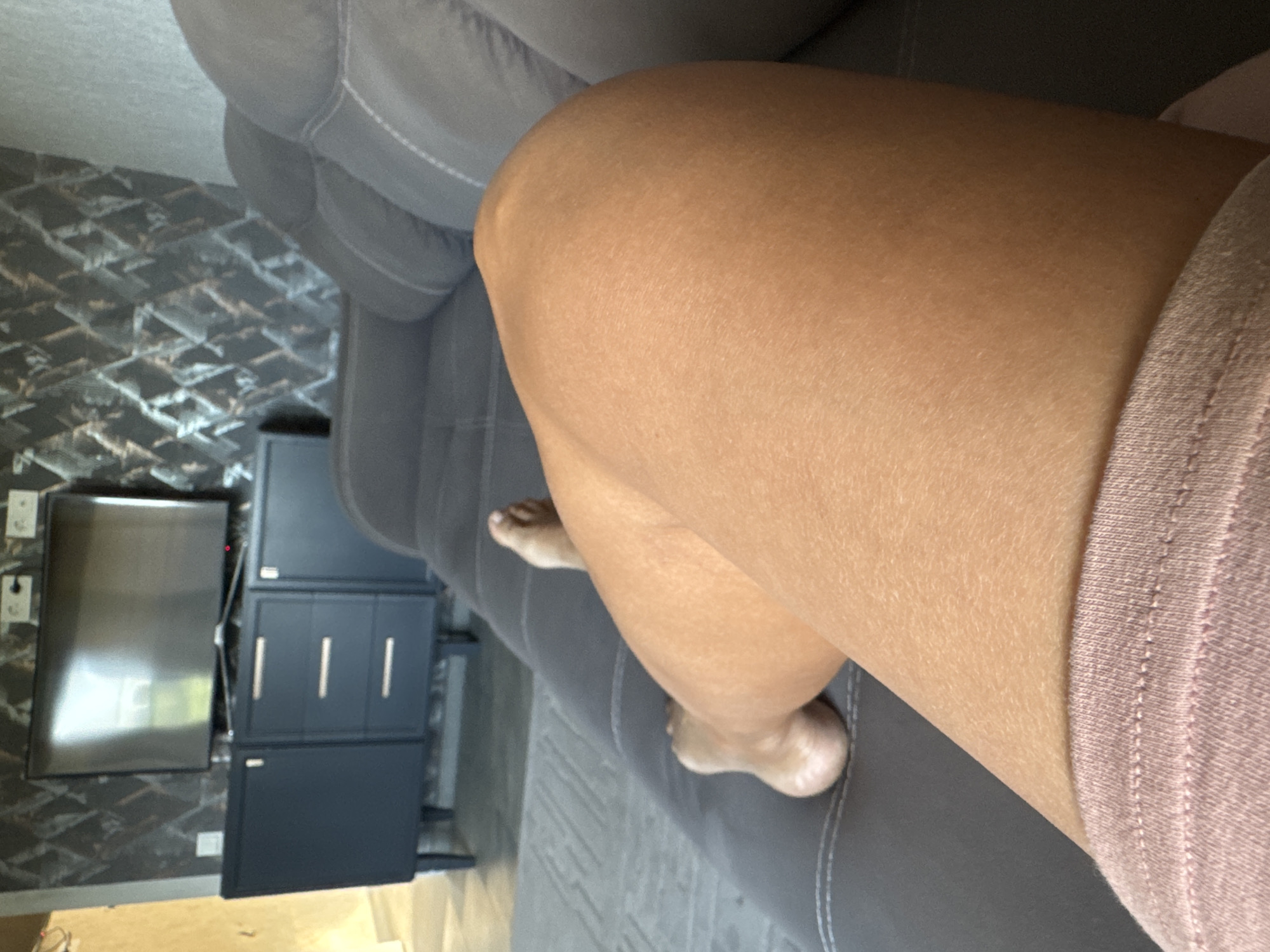 The image shows a person's legs resting on a gray sofa in a living room. The person is wearing pink shorts. In the background, there is a television mounted on a dark blue cabinet against a wall with patterned wallpaper. The location of Солонешное, Russia is not discernible from the image.
