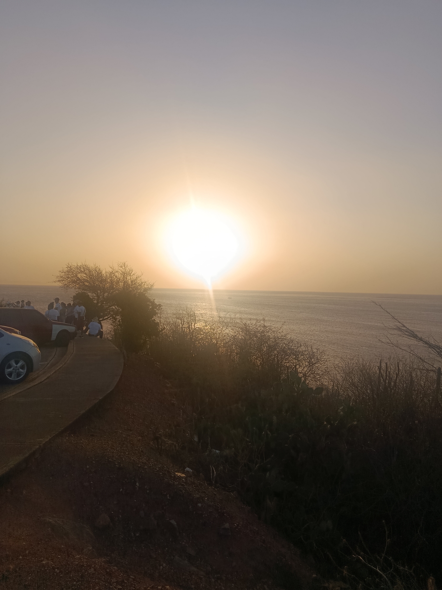 The image captures a sunset scene overlooking the ocean from Juan Griego, Venezuela. A group of people is gathered near parked cars along a curved concrete path. The sun is setting over the water, creating a bright glare on the surface of the sea. Vegetation covers the hillside in the foreground.