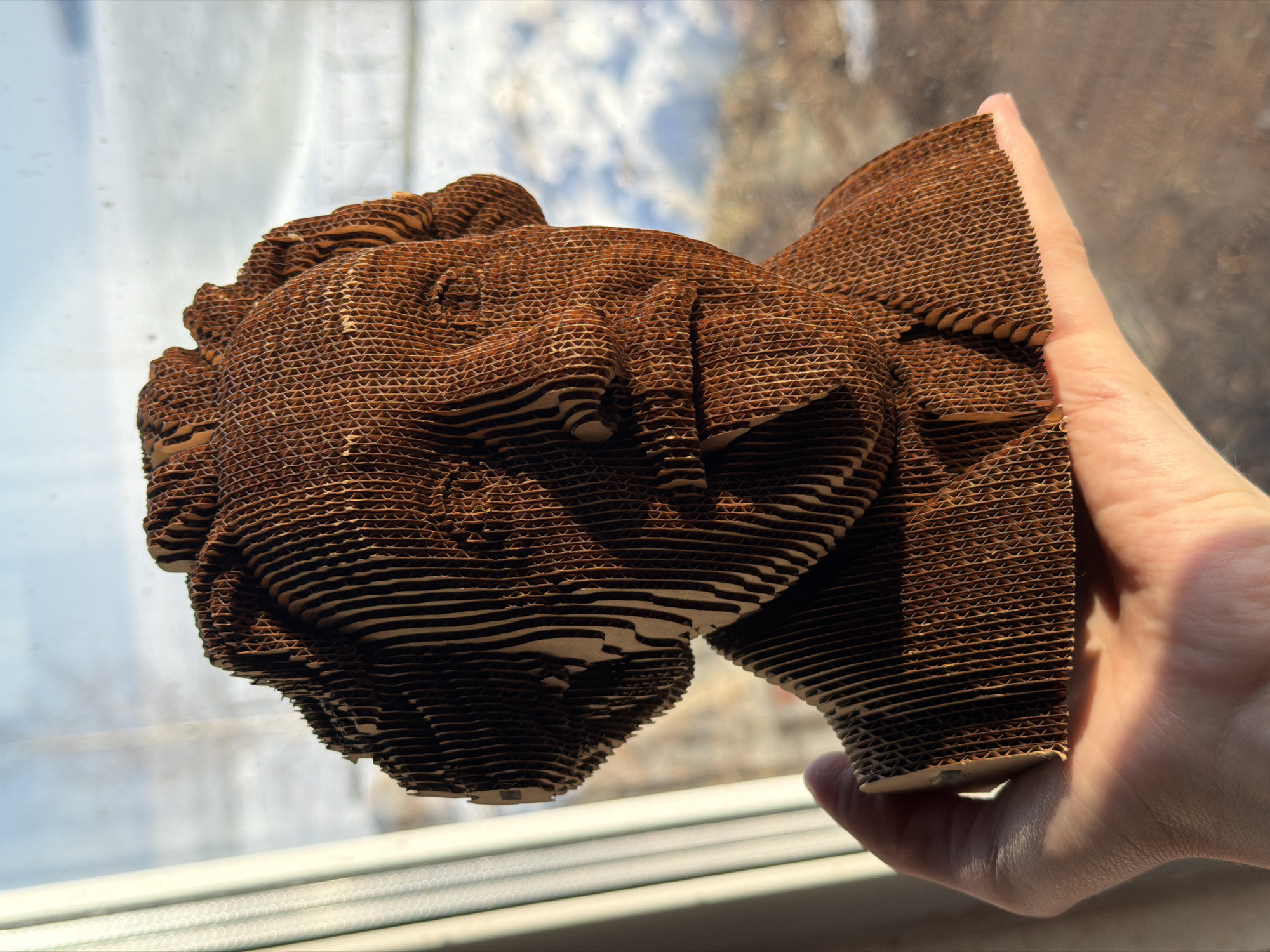 A forensic examination of the image reveals a close-up, waist-level shot taken indoors, looking out through a window onto a wintry landscape in the city of Chaikovsky, Russia. The central subject is a three-dimensional bust, approximately head-and-shoulder size, constructed from numerous horizontally layered pieces of corrugated cardboard. The bust depicts the likeness of a man, strongly resembling composer Pyotr Ilyich Tchaikovsky, with details such as facial features, hair, and collar discernible through the layering.
A human hand, consistent with an adult's, supports the base of the bust from below. The skin tone of the hand is fair, and sunlight casts distinct shadows across the palm and fingers. The corrugated edges of the cardboard layers are prominent, creating a textured, ridged appearance on the bust's surface.
Through the window in the background, a blurred outdoor scene is visible. This includes a vast expanse of light-colored, possibly snow or ice-covered ground, indicative of a frozen body of water or landscape. Above this, a clear, pale blue sky with minimal cloud cover is present. A vertical, light-colored curtain or blind is partially visible on the far left, bordering the window. Strong natural sunlight illuminates the scene from the right, highlighting the cardboard's texture and casting shadows that define the bust's contours and the hand's form. The overall impression is one of a bright, cold day.