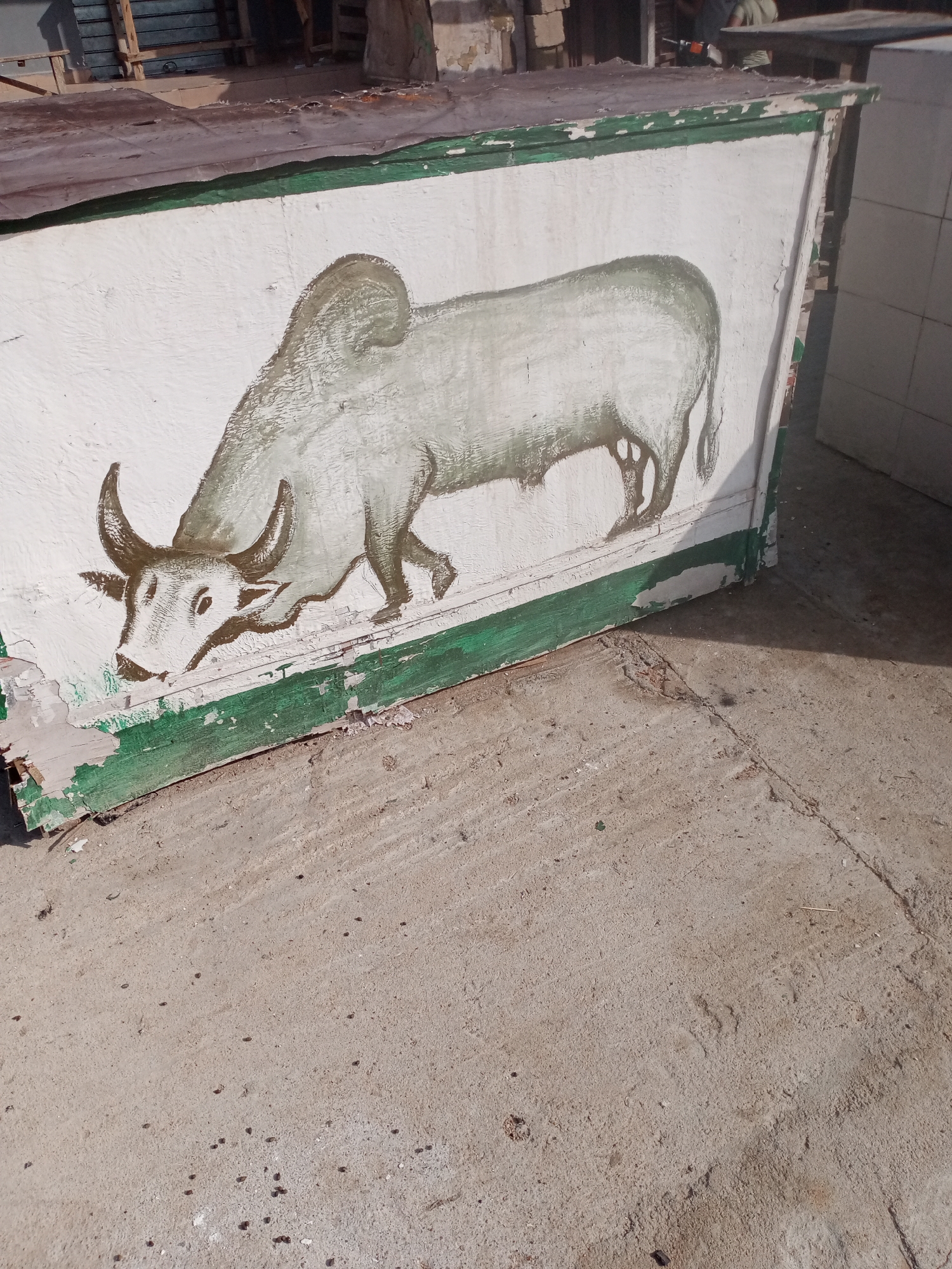 The image shows a painted depiction of a bull on the side of a structure. The painting is in a greyish-brown color and features the bull with its head lowered, as if grazing or drinking. The structure itself appears to be made of wood and is painted white with a green border along the bottom. The paint is weathered and peeling in several places. The ground in front of the structure is concrete and appears to be outdoors, with some debris scattered on it. In the background, there are glimpses of other structures and possibly some people, but they are out of focus and not clearly discernible. The scene is lit by bright sunlight, casting shadows. The overall impression is one of an outdoor setting, possibly a market stall or a workshop, in a somewhat rundown area. The presence of a bull painting might suggest a connection to agriculture or a local cultural motif. Without additional information, pinpointing the exact location in Jalingo, Nigeria is not possible. However, the scene is consistent with an urban or peri-urban environment in Nigeria.