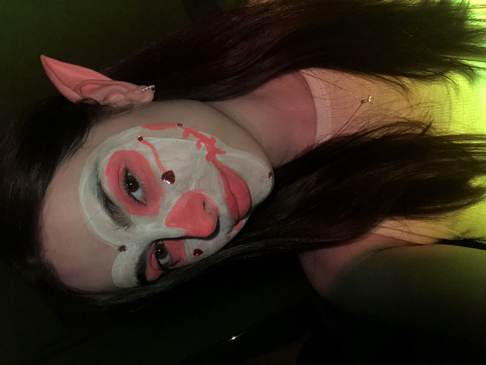 A close-up shot captures a person, likely a young woman, wearing elaborate face paint and prosthetic ears. She has long, dark hair, partially obscuring a light-colored top and a delicate necklace with a small charm. Her face is adorned with white face paint as a base, overlaid with striking pink patterns, including what resembles a pig-like snout and decorative elements around her eyes and cheeks. Small, red, heart-shaped gemstones or rhinestones are strategically placed on her face paint. Pink, pointed elf-like ears, decorated with tiny rhinestones, are visible on the side of her head. The person's eyes gaze directly at the viewer. The background is dimly lit with a vibrant artificial green and yellow glow, suggesting an indoor setting such as a party, costume event, or artistic performance. No text is visible in the image. The setting is likely an event taking place in Noviy, Russia.