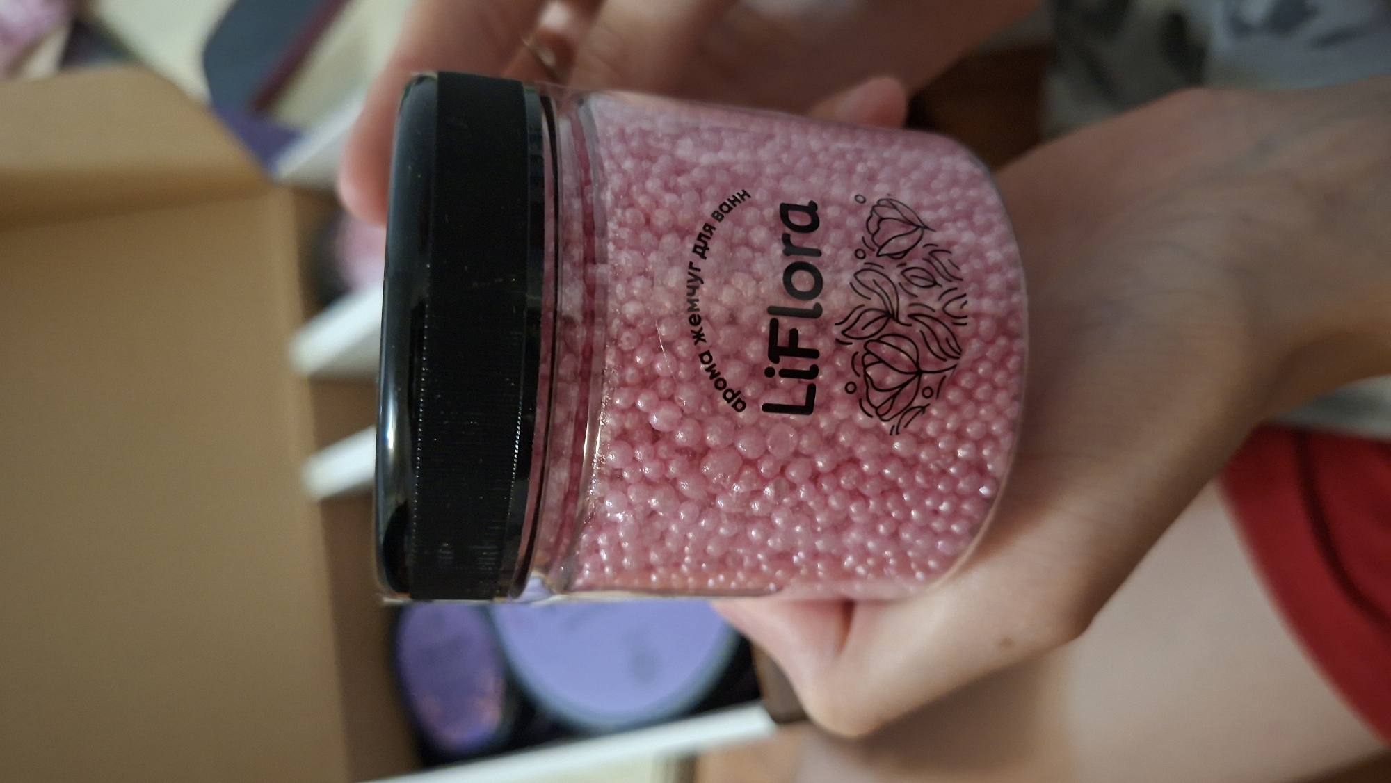 A person is holding a clear plastic jar filled with small, pink, pearl-like beads, which appear to be bath beads or aromatherapy pearls. The jar has a black screw-on lid and a label with the brand name "LiFlora" and some floral graphics. The text on the label is in Russian and says "арома жемчуг для ванн" which translates to "aroma pearl for bath". In the background, out of focus, are other items in containers, possibly part of a set or product display. The scene is likely indoors, possibly in a home or retail environment. No specific location in Забалоцкі сельскі Савет, Belarus can be determined from the image.