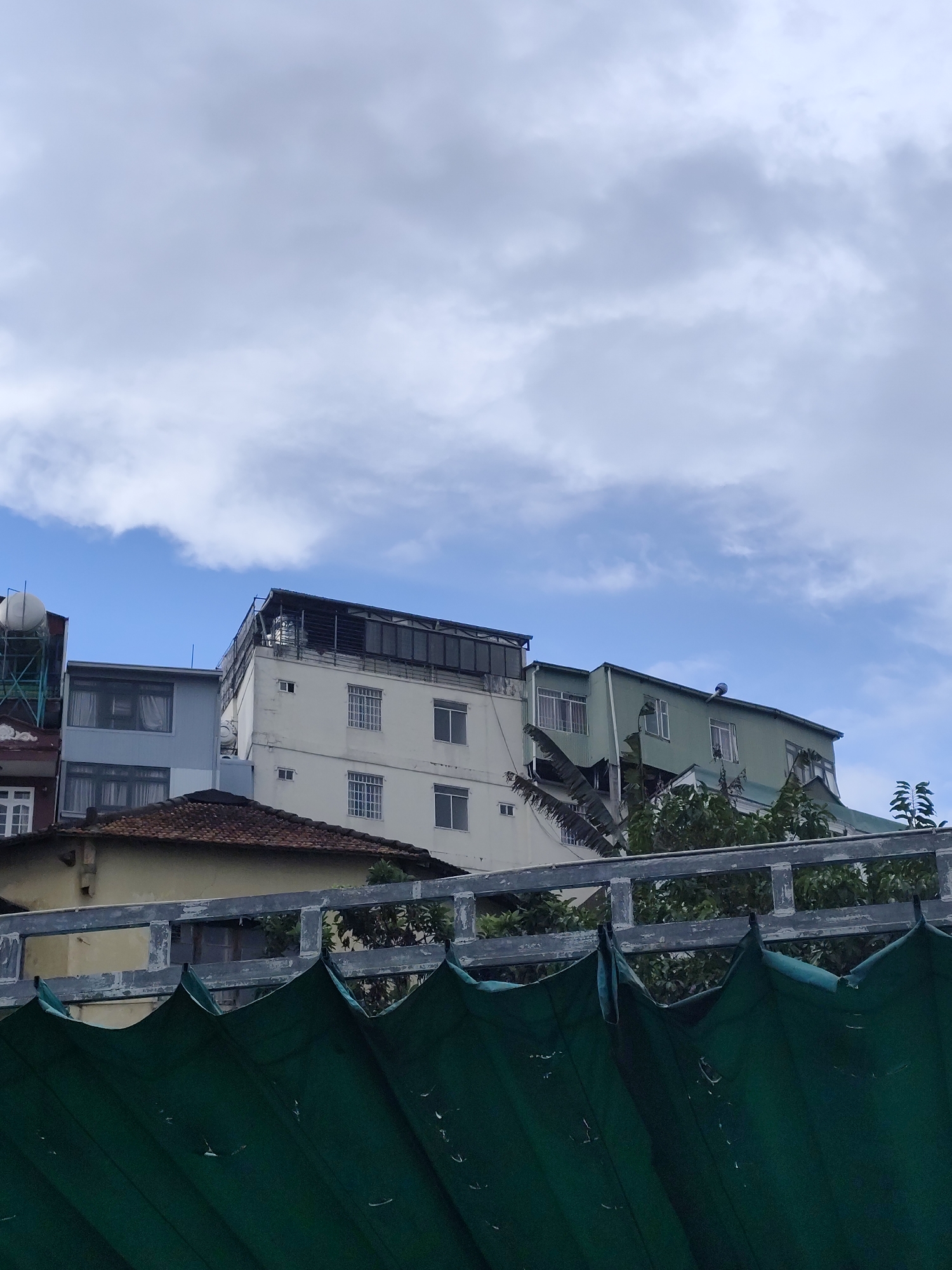 The image depicts an urban scene in Da Lat, Vietnam, characterized by multi-story buildings under a cloudy sky. In the foreground, a large, dark green tarp is draped, creating a textured barrier with visible pleats and some wear. Behind the tarp, a weathered gray metal railing with square openings separates the viewer from the buildings. Several buildings are visible, with varying architectural styles and colors. To the left, a light blue building with multiple windows is partially obscured. Next to it, a white building with numerous square windows and a darker rooftop structure stands prominently. To the right, a light green building with a sloping roof and windows is visible, with trees and foliage peeking through and around it. The overall impression is one of urban density and a somewhat overcast day in the city.