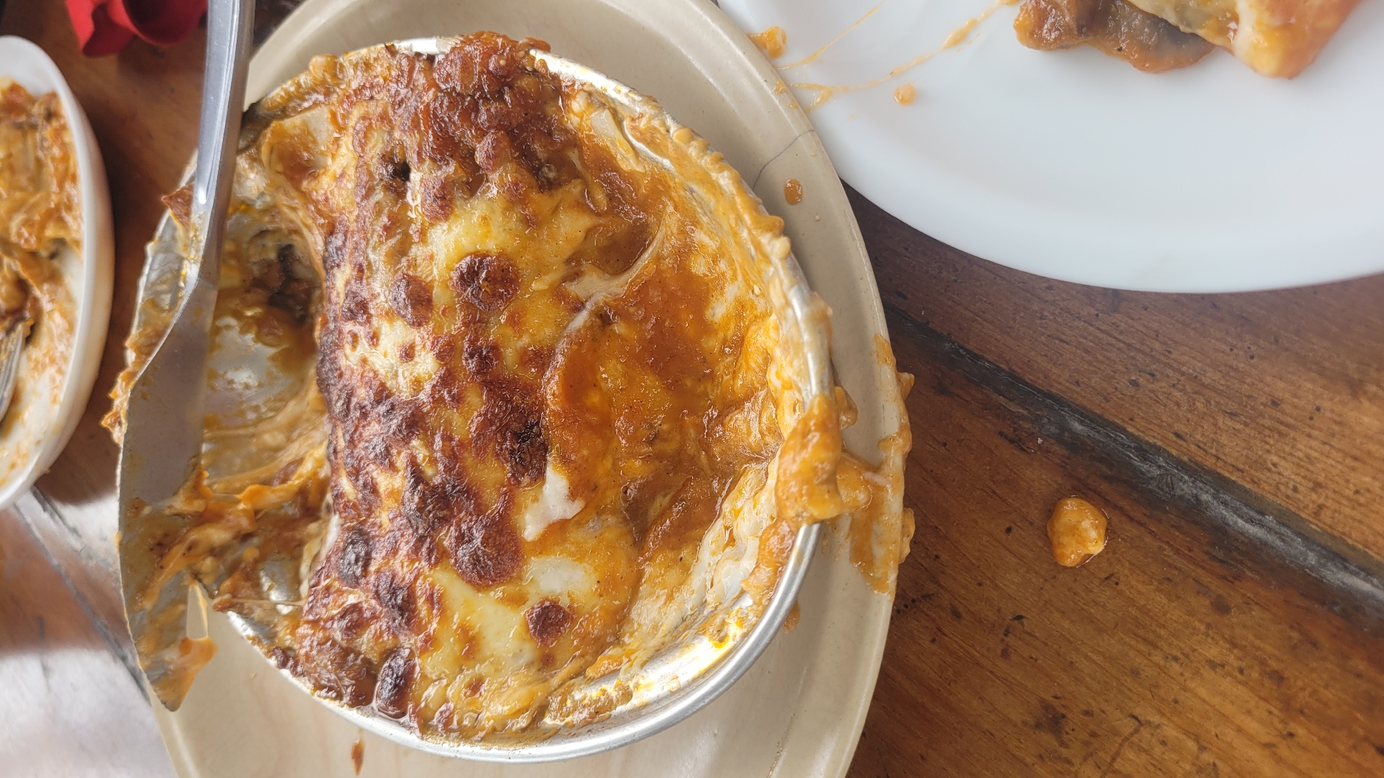 The image shows a close-up, overhead view of a serving of baked pasta, likely lasagna, in a round aluminum foil pan. The top layer is a bubbly, golden-brown melted cheese with some darker, crisped edges. A metal utensil, possibly a knife or spatula, is inserted into the dish, lifting a portion of the pasta and revealing layers of sauce and meat underneath. The pan rests on a light-colored plate, which is on a wooden table. In the background, another white plate with some leftover food is visible, along with a hint of a red napkin. The overall scene suggests a meal in progress or recently finished. The location is identified as Vashisht, India, though no specific Vashisht landmarks are visible in the image.