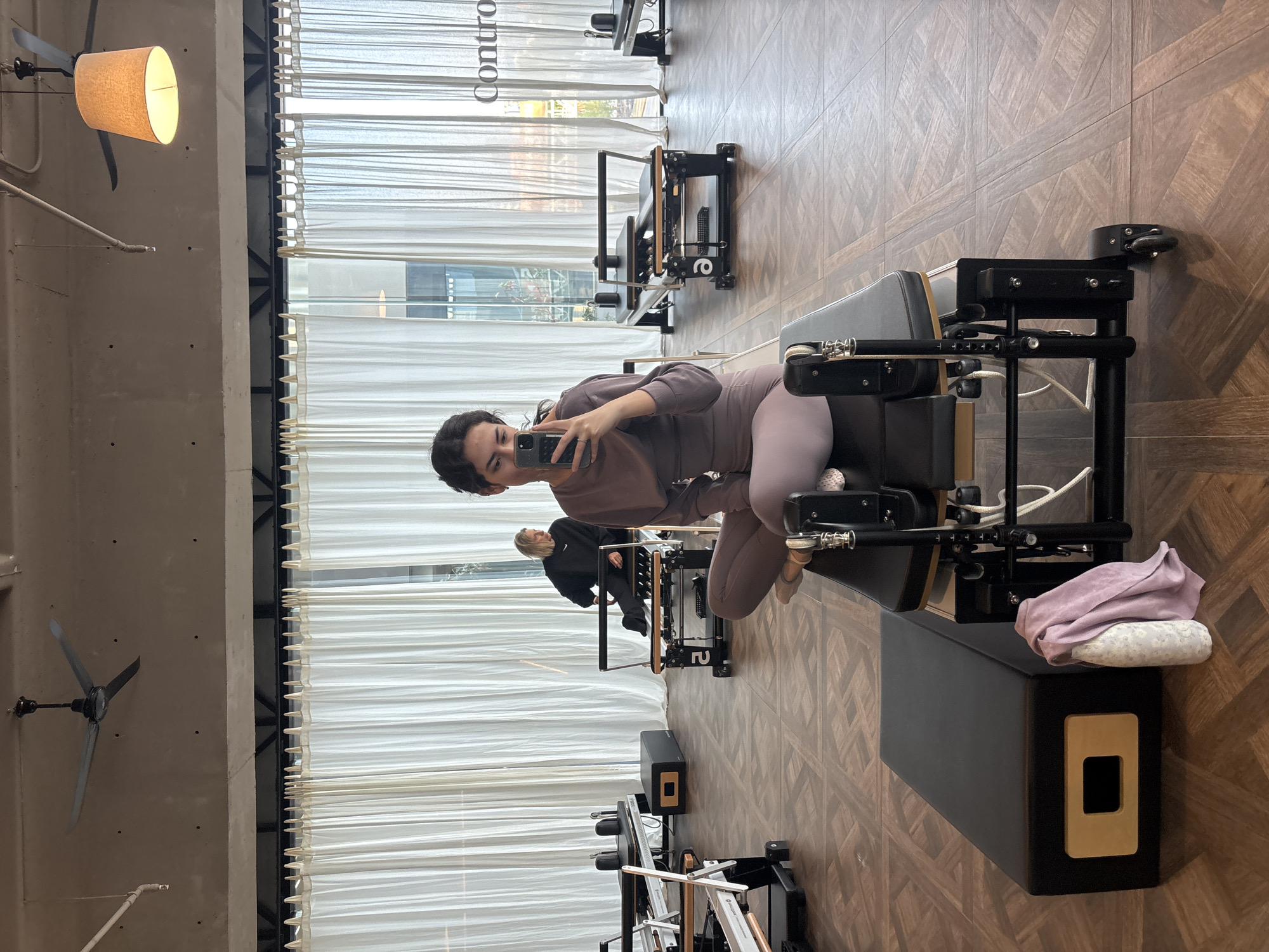 A woman is taking a selfie in a Pilates studio. She is sitting on a Pilates machine. Another person can be seen using a similar machine in the background. The studio has a wood floor and sheer curtains. A ceiling fan is visible in the corner of the room. The location is San Bernardino Tlaxcalancingo, Mexico.