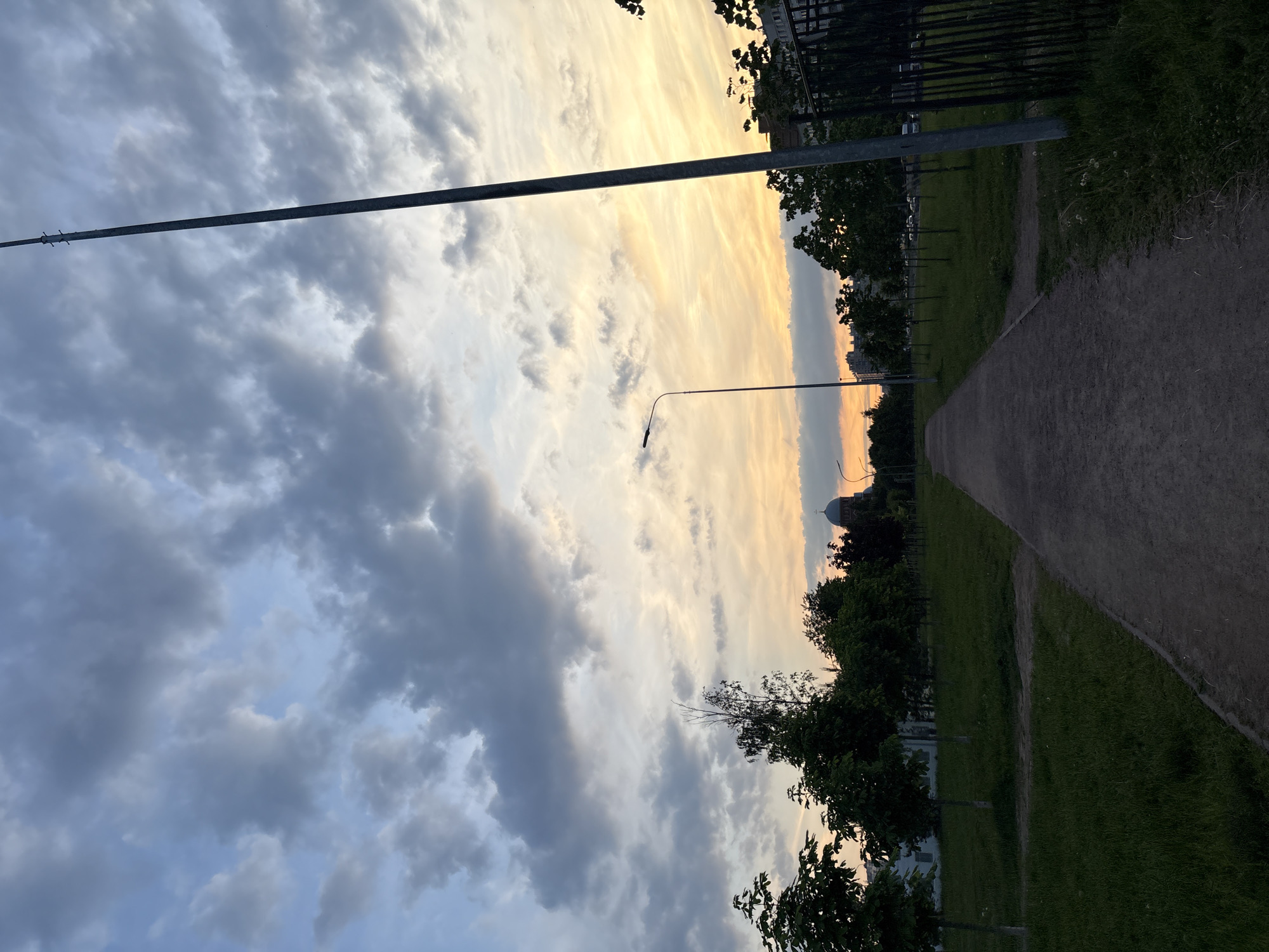 The image depicts a paved pathway leading through a park-like setting in Sinyavino, Russia. The pathway is flanked by green grass and scattered trees, some of which are young saplings. In the distance, across a line of trees, a building with a dome and a cross is visible, suggesting it might be a church or a similar structure. The sky above is filled with dramatic clouds, transitioning from a muted blue at the top to soft oranges and yellows near the horizon, indicative of either sunrise or sunset. A tall, slender lamppost stands prominently on the right side of the pathway, and another lamppost is visible further down the path. A dark metal fence runs along the right edge of the frame, partially obscured by grass and vegetation. The overall impression is a tranquil, urban park scene during twilight.