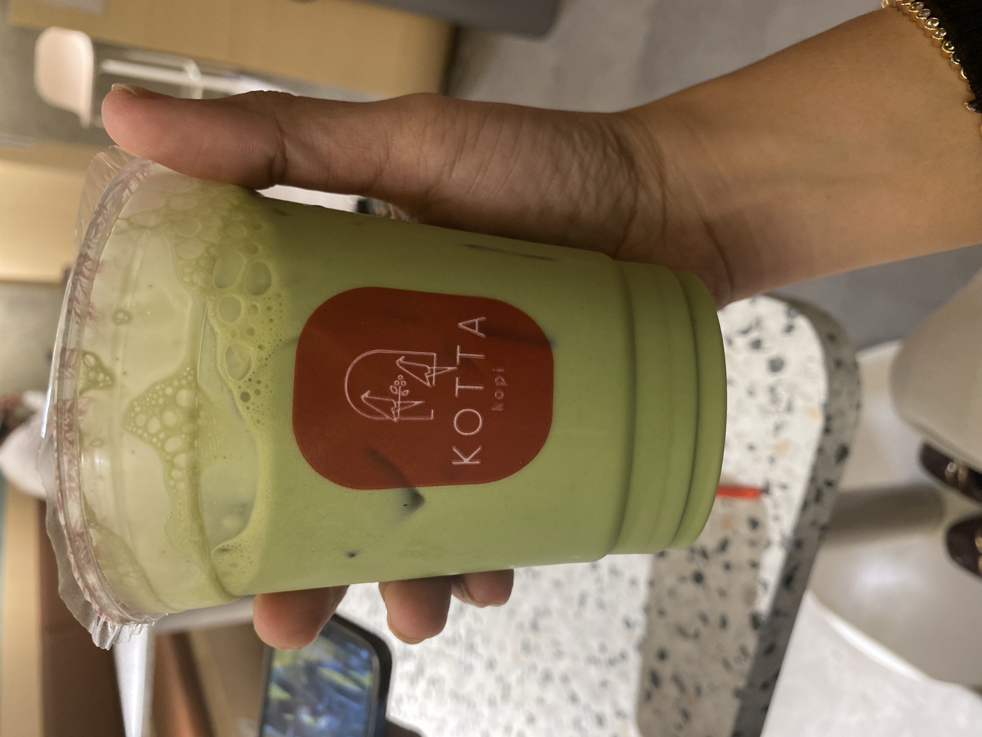 A close-up, eye-level shot captures a hand holding a clear plastic cup filled with a vibrant green beverage, likely a matcha latte, topped with a frothy layer. A circular, reddish-brown sticker with a white outline of a bird-like figure and the text "KOTTA kopi" is adhered to the cup. The background is blurred, suggesting an indoor setting. To the left, a portion of a mobile phone is visible. To the right, a person's arm, adorned with a gold bracelet, is also in view. In the distance, a white bar stool can be seen. The overall impression is a casual moment, possibly at a cafe, within the city of East Jakarta, Indonesia, given the branding on the cup.
