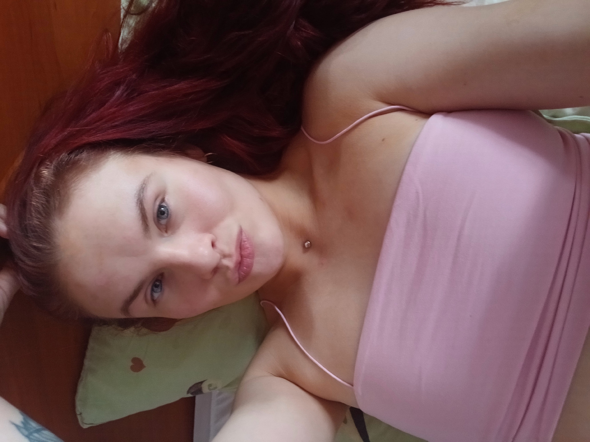 A fair-skinned woman with shoulder-length dark red hair and blue eyes is lying down on a green pillow patterned with red hearts. She is wearing a pink tube top with thin straps. A piercing is visible on her neck. The location is presumed to be indoors in an unknown location in Chaikovsky, Russia.