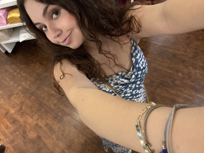 A brunette woman with curly hair poses for a selfie in what appears to be a store. She wears a blue and white patterned dress with a draped neckline and gold chain straps. On her left wrist are several bracelets. The background includes wooden flooring and a shelf with stacks of folded items in varying colors.
