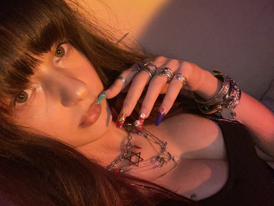 A woman with dark hair and bangs stares at the camera. Her hand, adorned with multiple rings and long, decorated nails, rests near her face. She wears several bracelets and layered necklaces, one with a Star of David. The lighting is warm and shadowed, revealing her cleavage. The overall impression is bold and sensual.