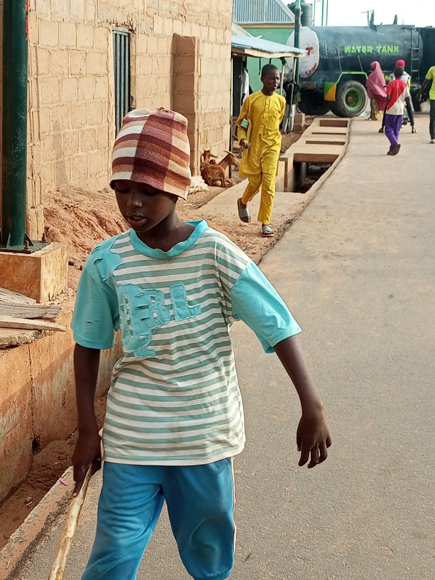 In Tempure, Nigeria, a young Black boy wearing a striped beanie and a light blue and white striped t-shirt walks down a paved road, holding a wooden stick. He is in the foreground, and the camera is at his eye level. To his left, a rough brick building stands, with a green metal pole and a brown dirt pile next to it. In the background, several people are walking near a large black water tank truck. One person in a yellow outfit walks in the same direction as the boy in the foreground. Another person in a pink headscarf and purple pants walks away from the camera. The environment appears to be outdoors in a rural or semi-urban setting.