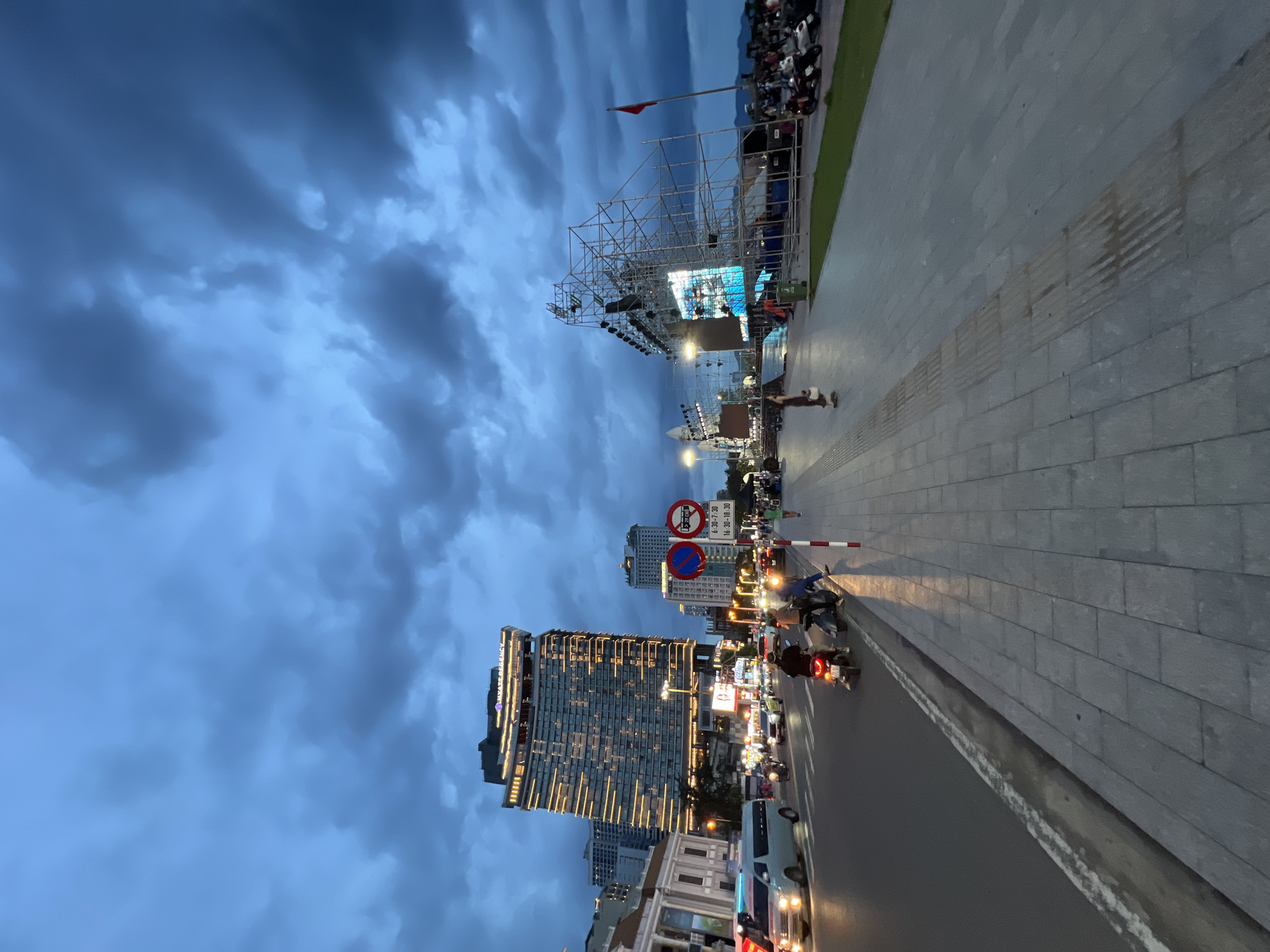 The image depicts a bustling street scene in Nha Trang, Vietnam, at dusk. A large, illuminated hotel, identified as the Hyatt Regency Nha Trang, dominates the skyline. In the foreground, a wide pedestrian walkway runs alongside a busy road where cars and motorcycles are in motion. A large temporary stage structure with a screen is set up on the right side of the walkway, indicating an ongoing event or performance. Several people are visible walking on the sidewalk and riding motorcycles on the street. The sky is filled with dramatic, dark clouds, suggesting impending weather. Traffic signs, including a no-entry sign and a timed parking restriction sign, are visible along the road. The overall atmosphere is one of urban activity and a sense of anticipation, perhaps for the event at the stage.