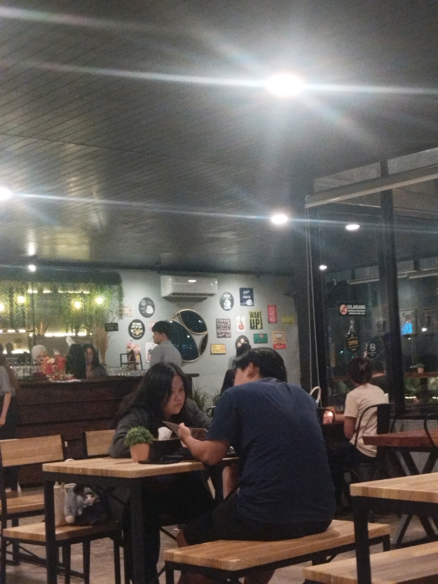 The image depicts the interior of a cafe. In the foreground, a young woman with long dark hair is seated at a table, looking down at a tablet. She is wearing a dark sweater. A man in a dark blue t-shirt is seated next to her, also looking at the tablet. Their table is made of wood with black metal legs, and two wooden chairs are visible. Under the table, a stuffed animal is placed inside a bag.
The cafe's walls are decorated with various framed signs, including one that says "WAKE UP!". There are also hanging plants and a wall-mounted air conditioning unit. In the background, other patrons and staff are visible. Through a glass partition, a person in a light-colored shirt is seated at another table. The ceiling is grey and appears to be paneled with metal or wood.
While the specific location in Sungailiat, Indonesia, cannot be determined from the image alone, the presence of Indonesian text on some signs (e.g., "DILARANG" meaning "forbidden") suggests the country. The overall atmosphere is casual and dimly lit, characteristic of a cafe.